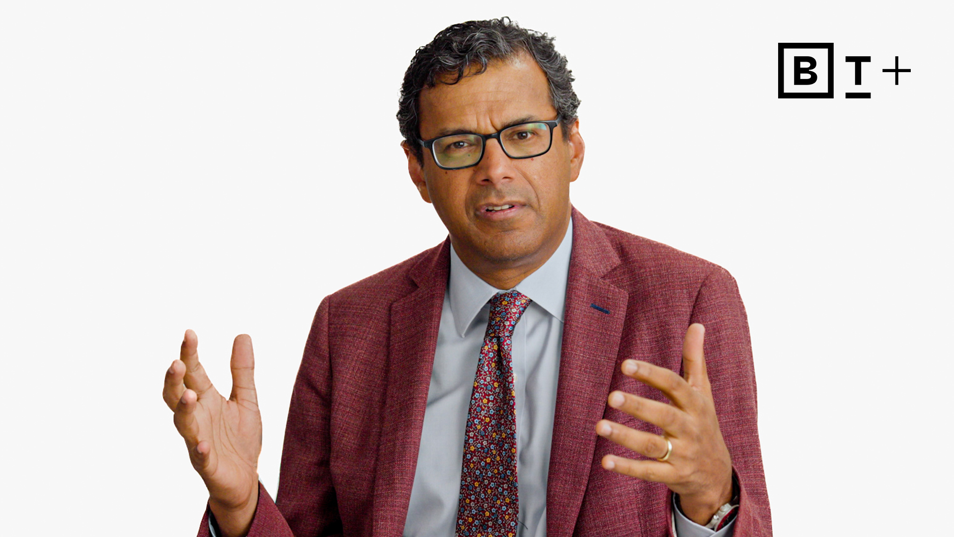 A man in a red suit jacket and patterned tie gestures with both hands while speaking. The background is plain white with the "B T+" logo in the top right corner.