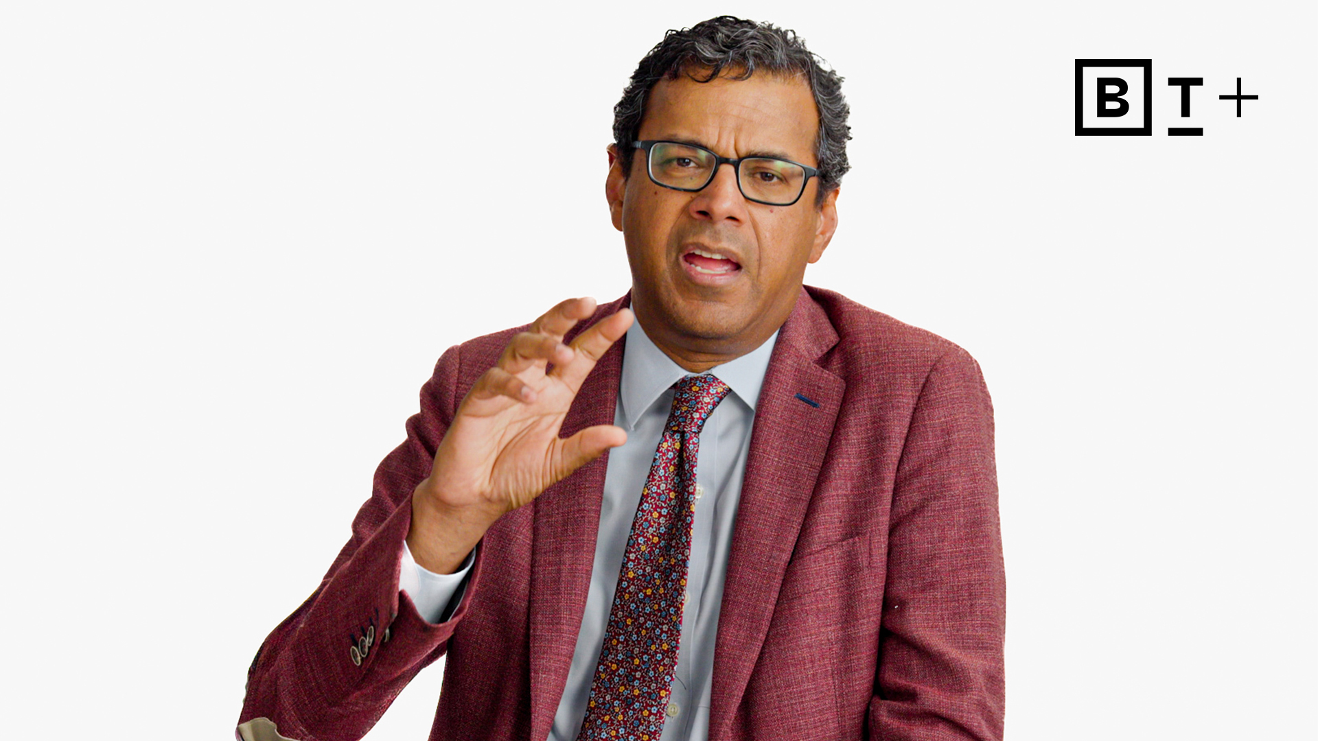 A man in a suit and tie gestures with his hand while speaking, sitting against a plain white background with a "BT+" logo in the top right corner.
