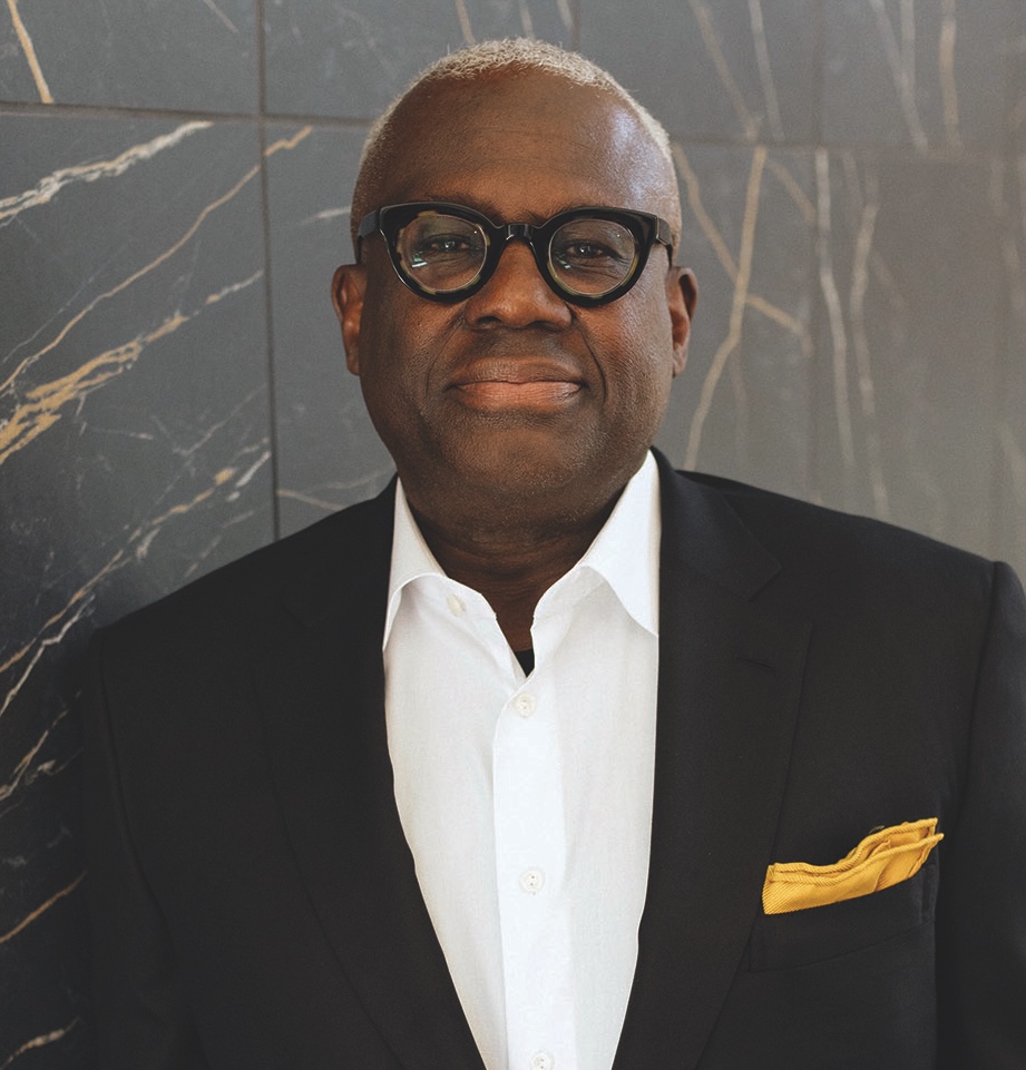 A man wearing glasses, a dark suit with a yellow pocket square, and a white shirt stands in front of a marble wall.