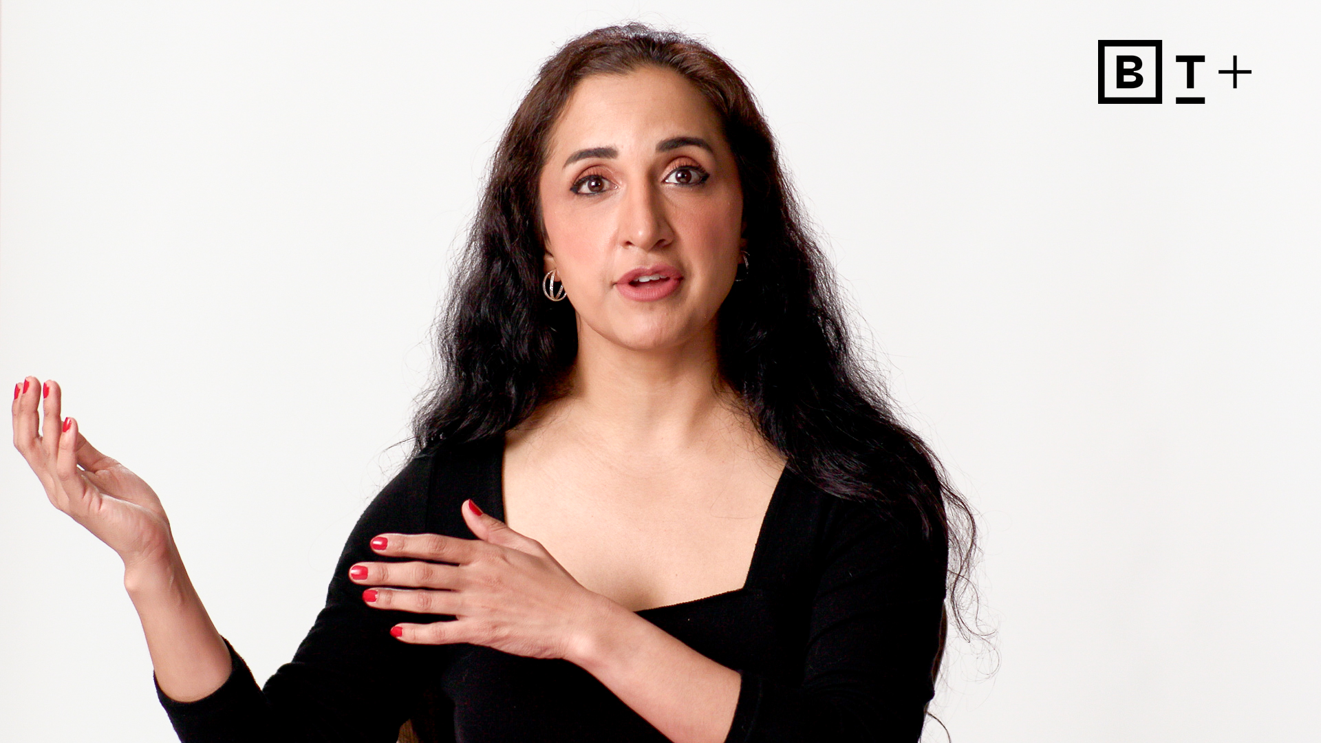A person with long dark hair gestures with one hand and touches their shoulder with the other, standing against a plain white background with a "BT+" logo in the top right corner.