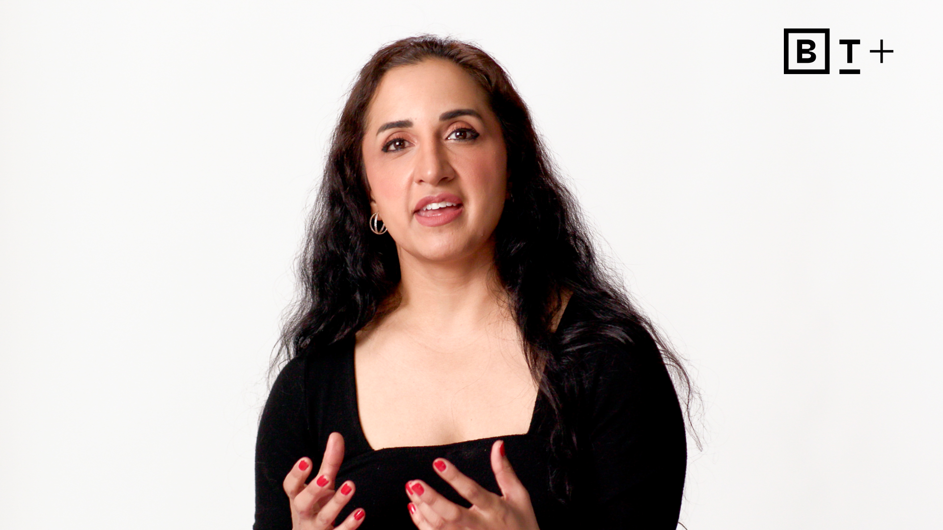 A woman with long, dark hair and a black top speaks while gesturing with her hands against a white background. The BT+ logo is in the top right corner.