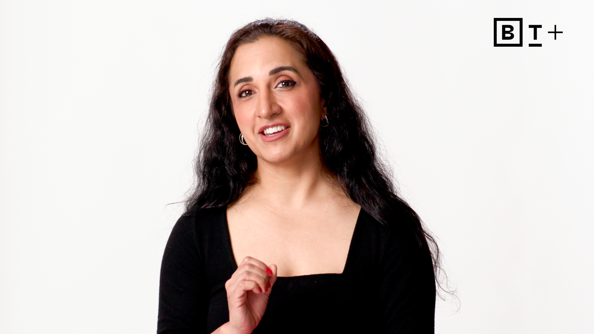 A woman with long dark hair, wearing a black top, speaks while gesturing with her hand. The background is white, with a "BT+" logo in the top right corner.