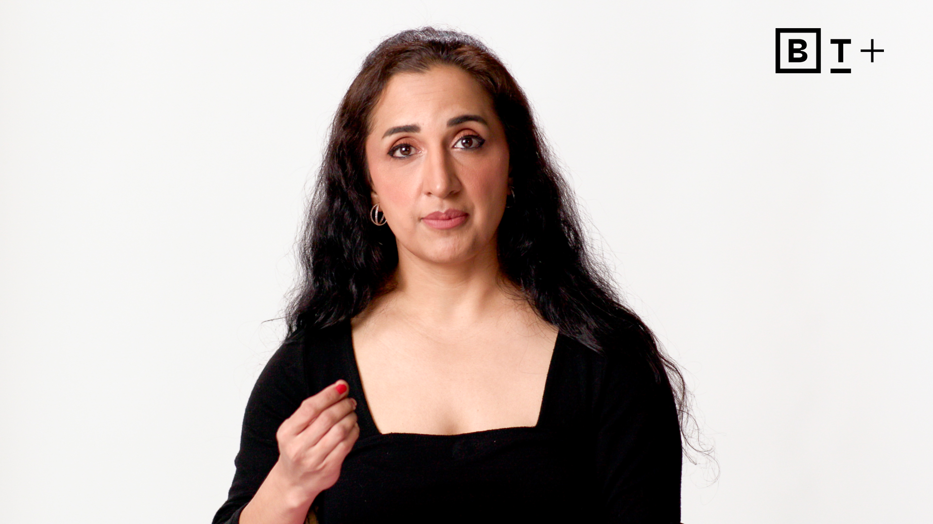 A woman with long dark hair in a black top gestures with her right hand against a plain white background. "B T +" logo appears in the top right corner.