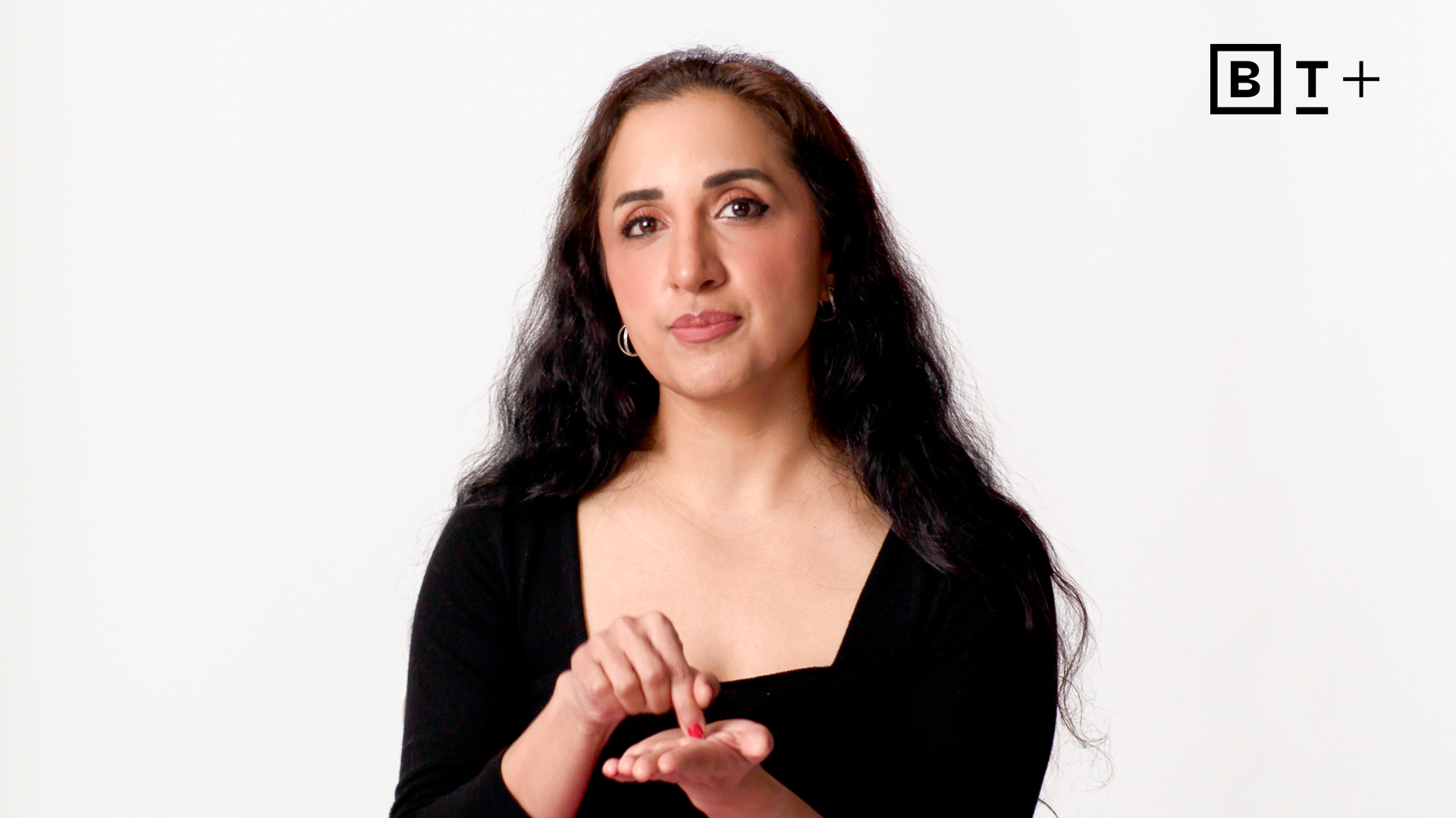 A woman with long dark hair wearing a black top gestures with her hands, standing against a plain white background with a "BT+" logo in the top right corner.