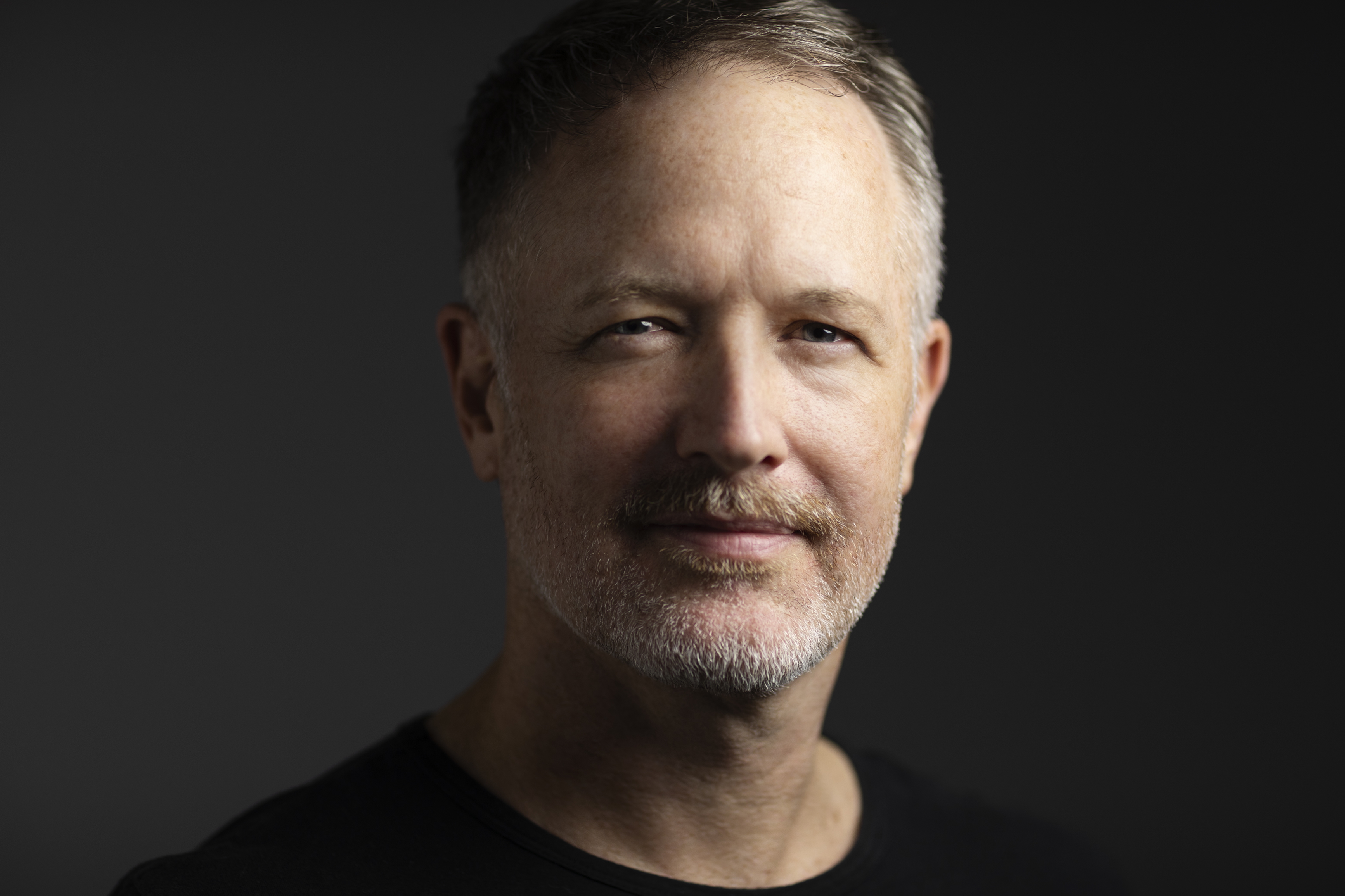 A middle-aged man with short hair and a trimmed beard, wearing a black shirt, is looking at the camera against a dark background.