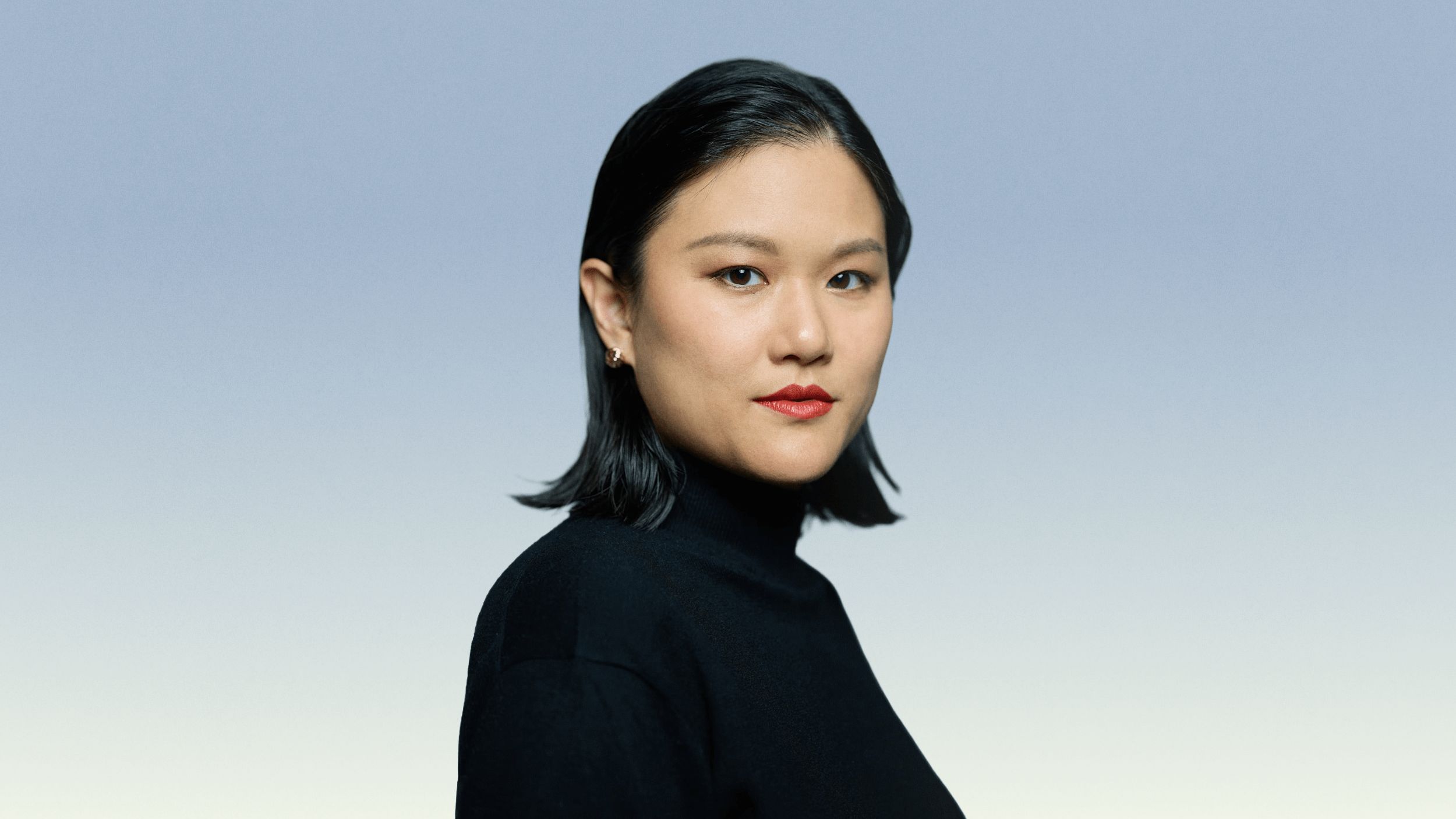 A woman with straight black hair, wearing a black turtleneck, poses against a blue-to-white gradient background.