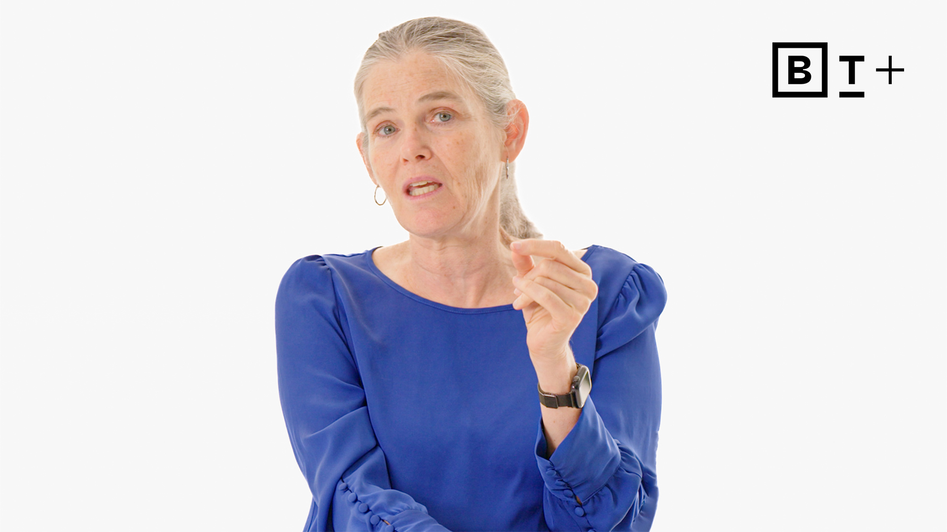 A woman with gray hair in a blue blouse speaks while gesturing with her hand. A "BT+" logo is in the top right corner on a plain white background.