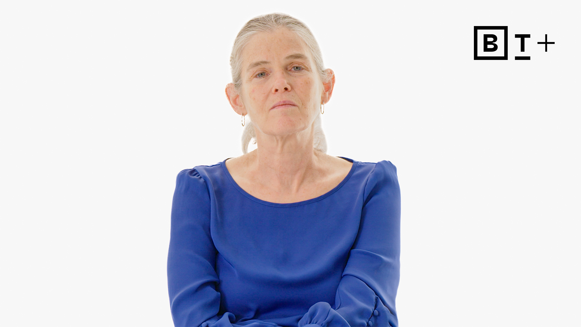 A woman with gray hair wearing a blue blouse sits against a plain white background with a "BT+" logo in the top right corner.