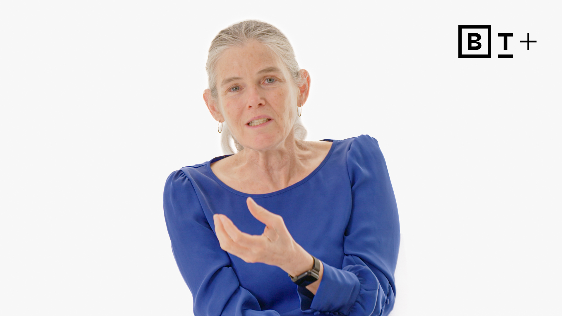 An older woman with gray hair, wearing a blue long-sleeve top, gestures while speaking against a plain white background with a "B T+" logo in the upper right corner.