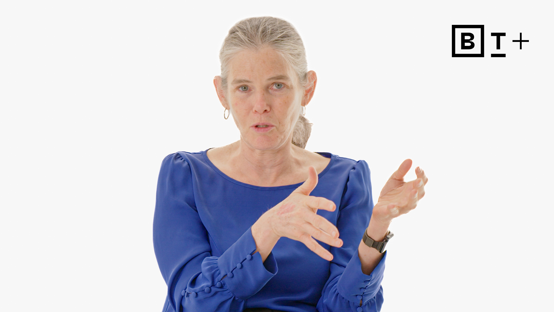 A woman with gray hair tied back, wearing a blue blouse, gestures with her hands while speaking. The background is white with a "BT+" logo in the top right corner.