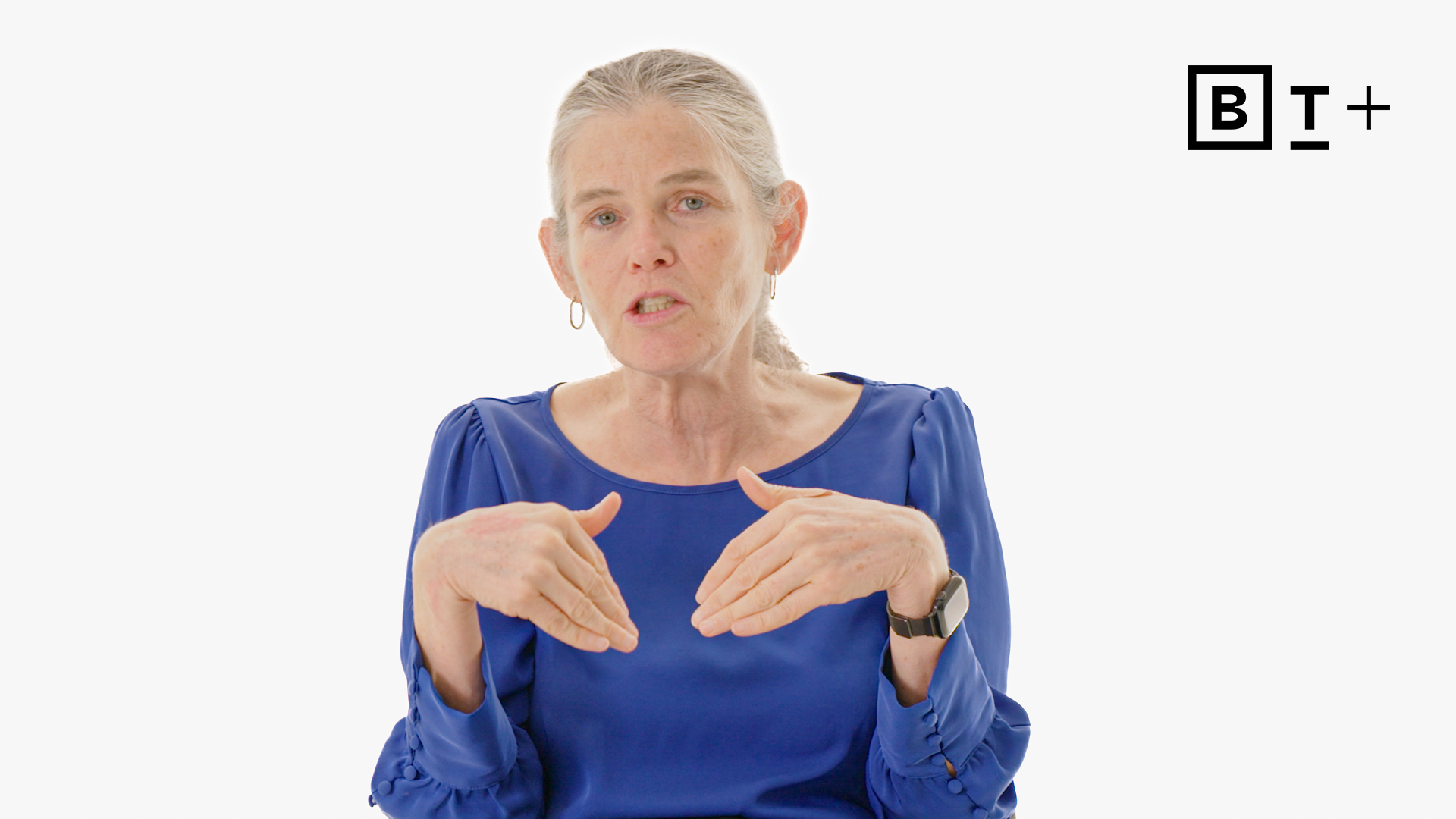 A woman with gray hair in a ponytail, wearing a blue blouse and watch, gestures with her hands. The background is white with a "B T +" logo in the top right corner.