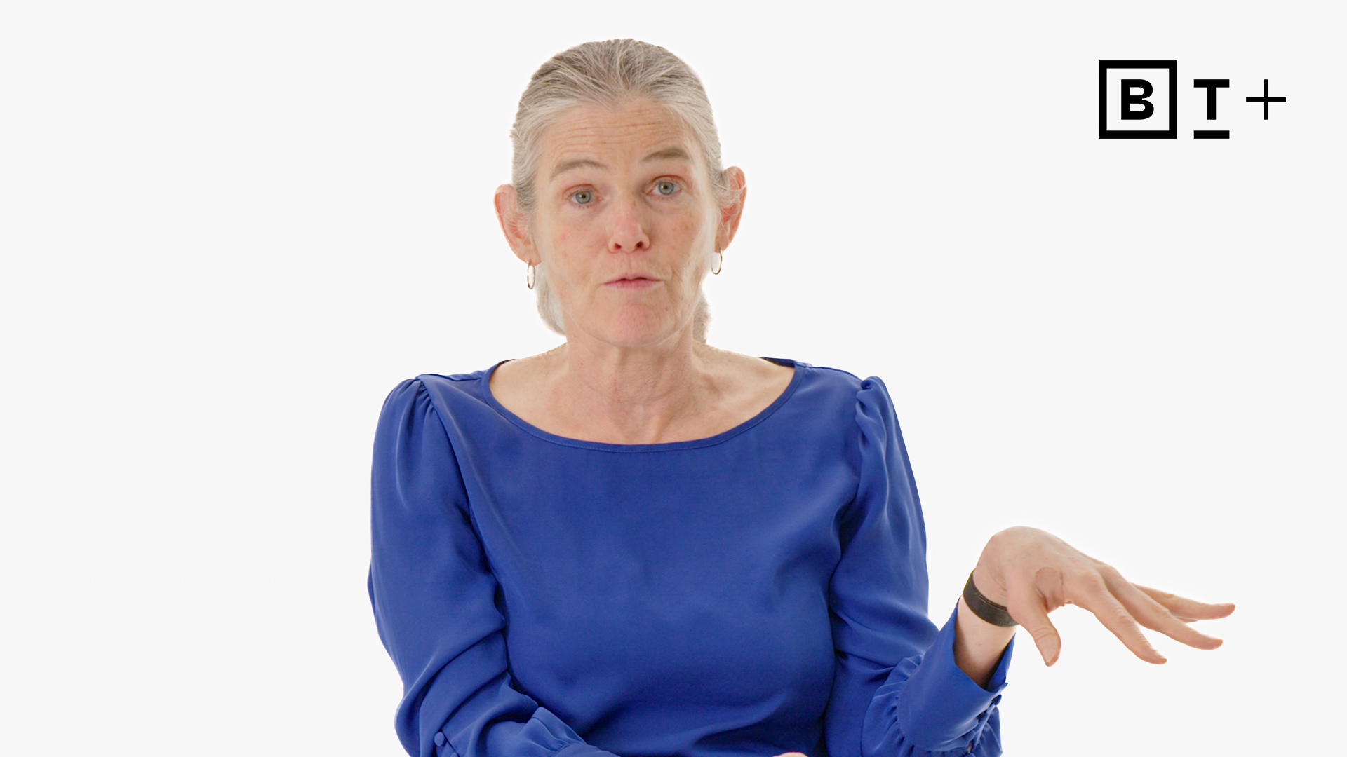 A woman with gray hair in a low ponytail, wearing a blue blouse, gestures with her hand while seated against a plain background with a "B T +" logo in the top right corner.