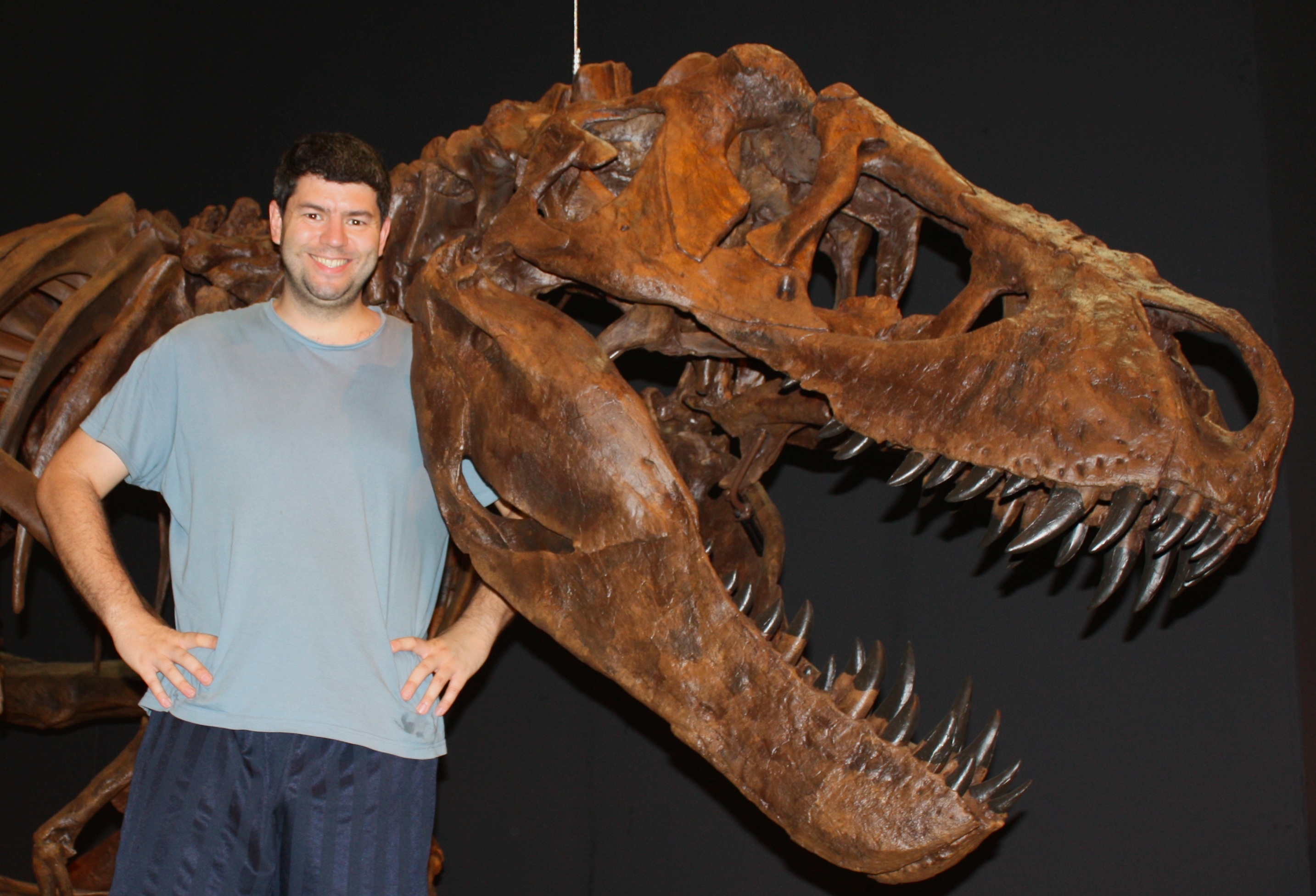 A man stands smiling next to a large dinosaur skeleton, likely a Tyrannosaurus rex, against a dark background.