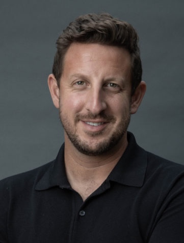 A man with short brown hair and a beard, wearing a black collared shirt, smiles at the camera against a plain gray background.