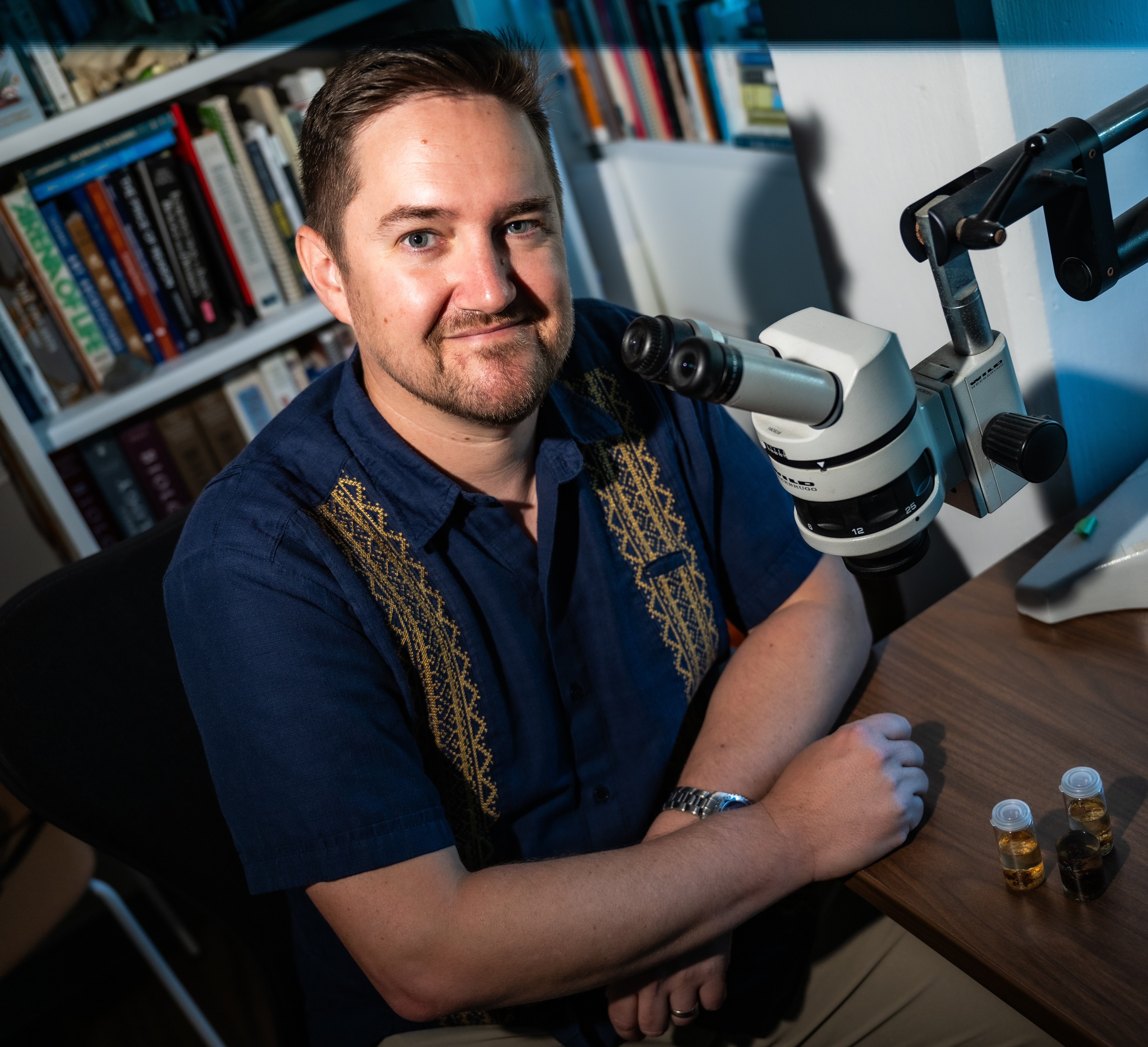 A man sits at a desk with a microscope, two small vials, and bookshelves in the background, looking at the camera and smiling.