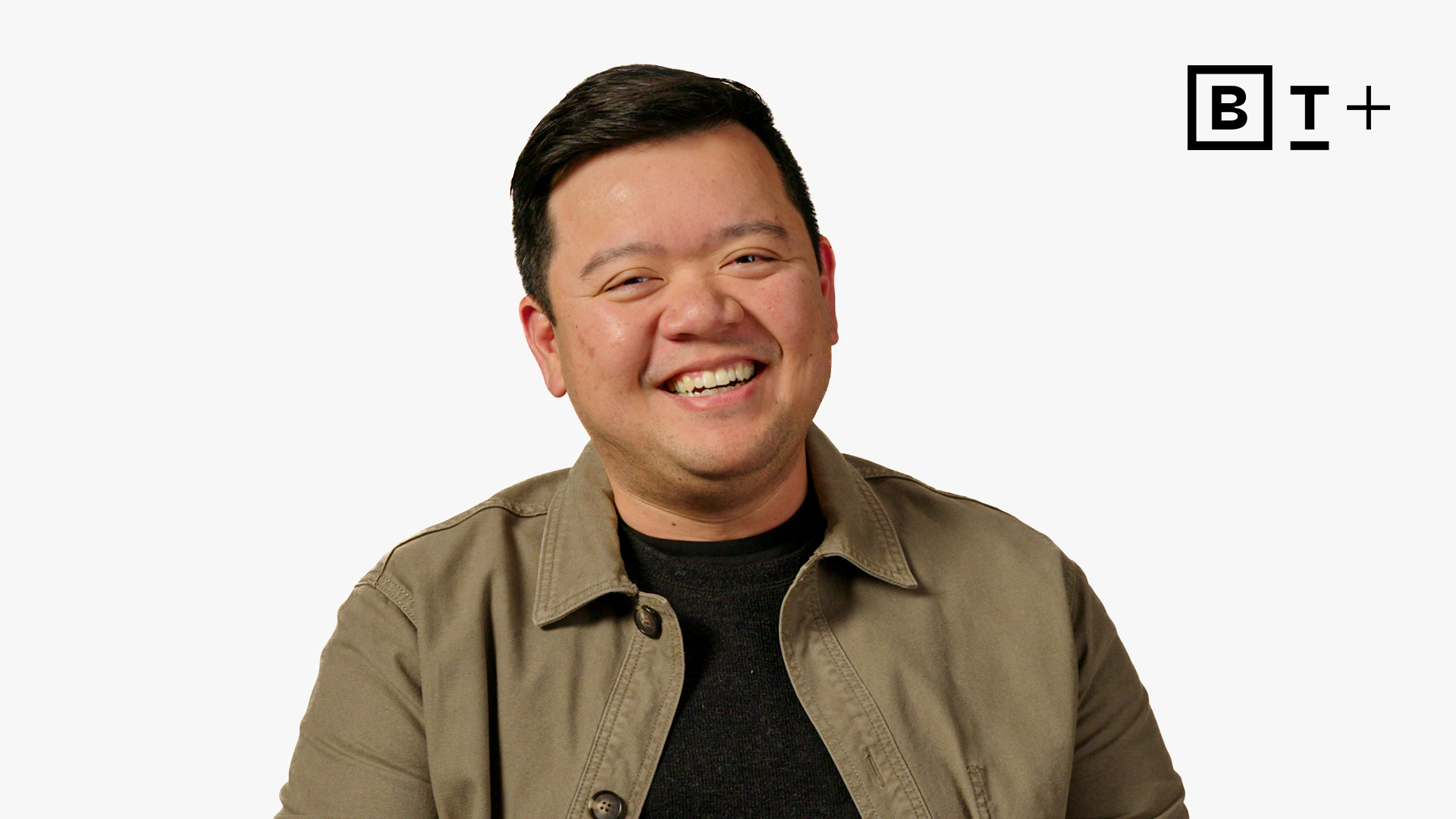 A man with short dark hair smiles while wearing a tan jacket over a dark shirt. The background is white with a "B+" logo in the top right corner.