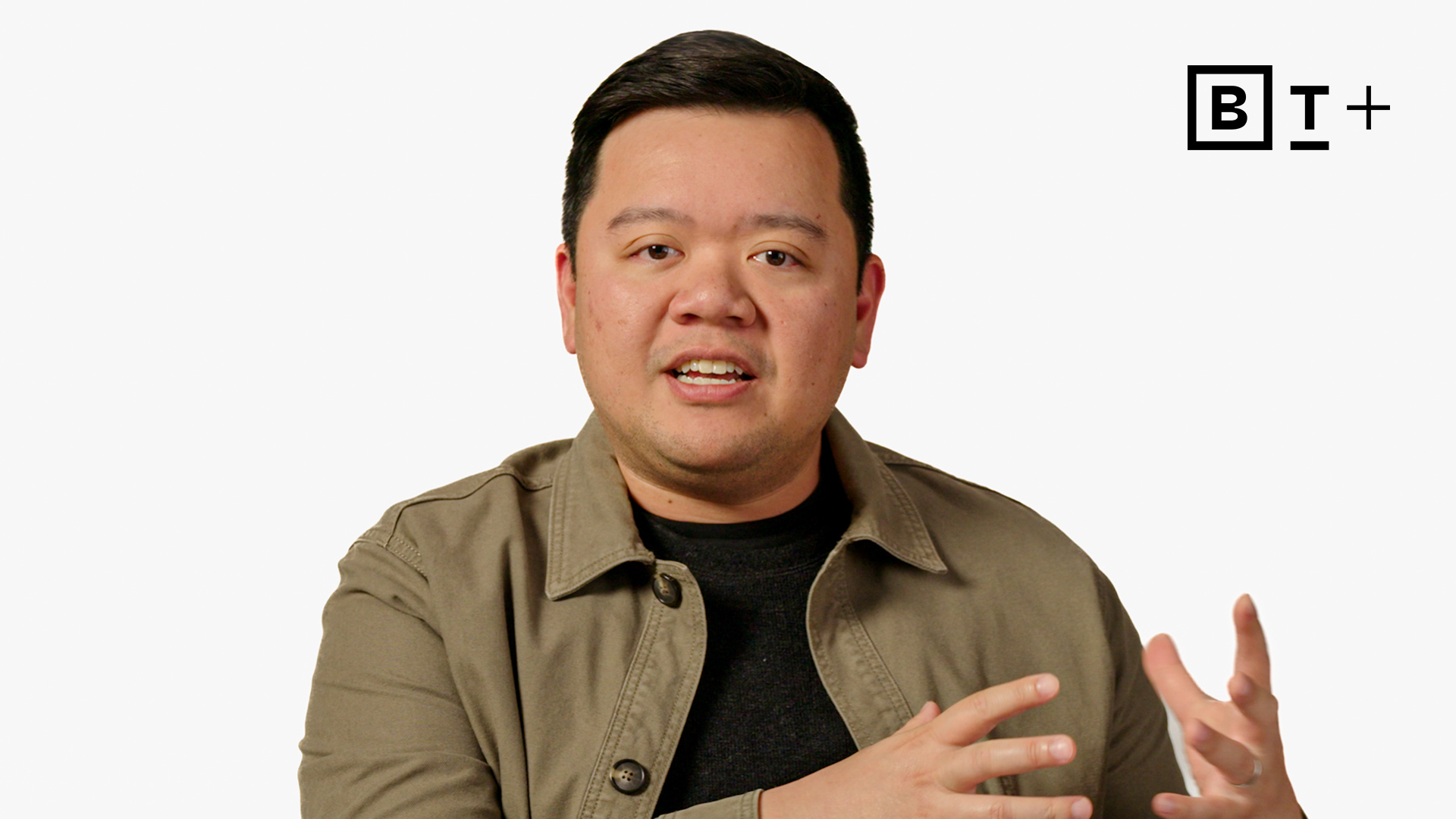 A person in a beige jacket gestures with hands while speaking against a plain white background with a "BT+" logo in the corner.