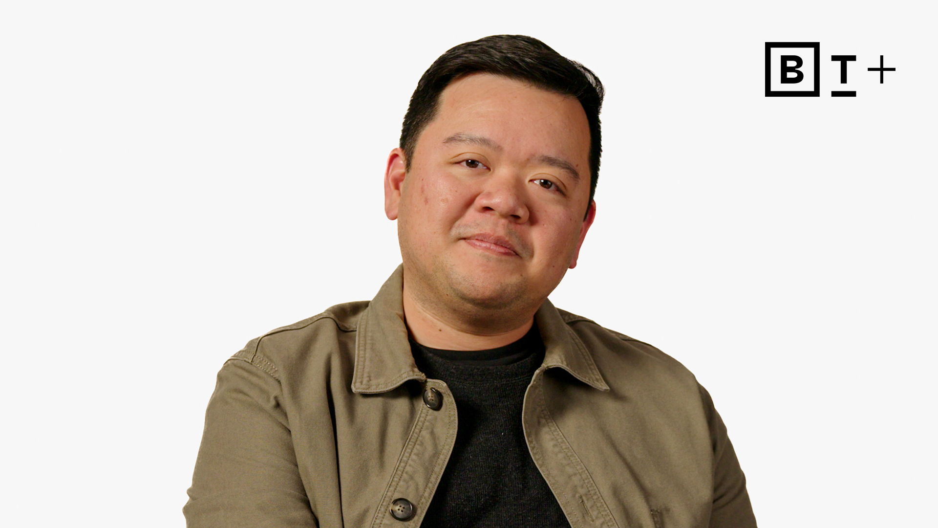 A man in a tan jacket and black shirt sits in front of a white background with a "B T +" logo in the top right corner.