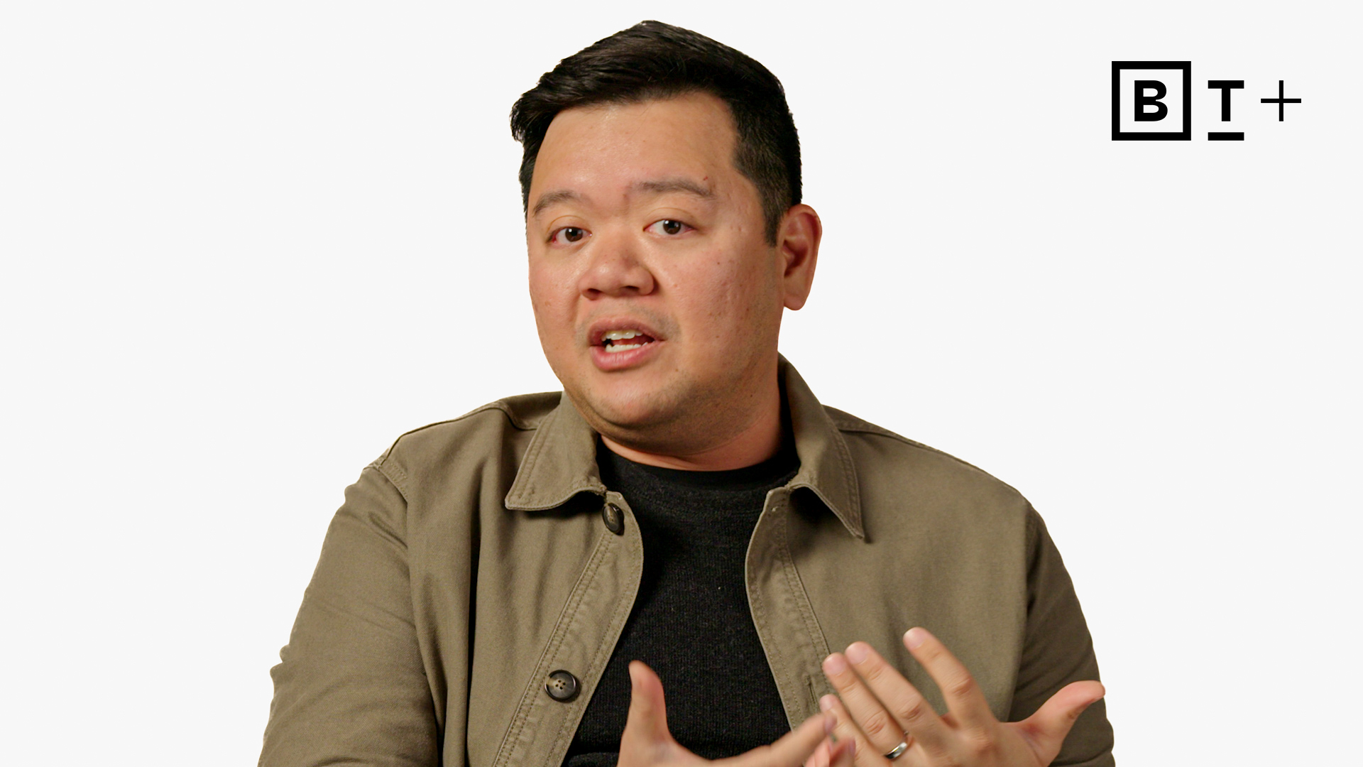 A man in a khaki jacket gestures with his hands while speaking, with a "BT+" logo in the top right corner on a plain white background.