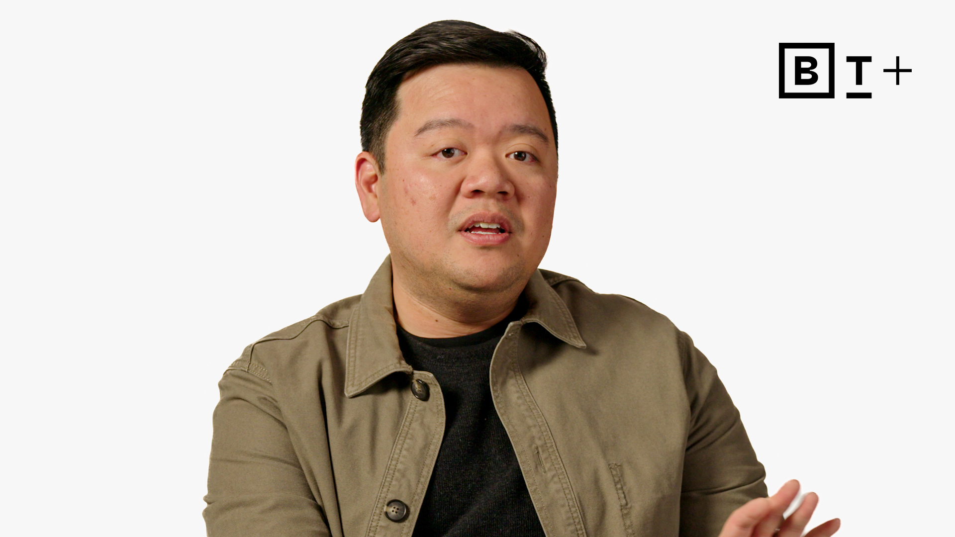 A man wearing a khaki jacket and black shirt speaks, gesturing with his hand. The background is plain white with a stylized "BT+" logo in the top right corner.