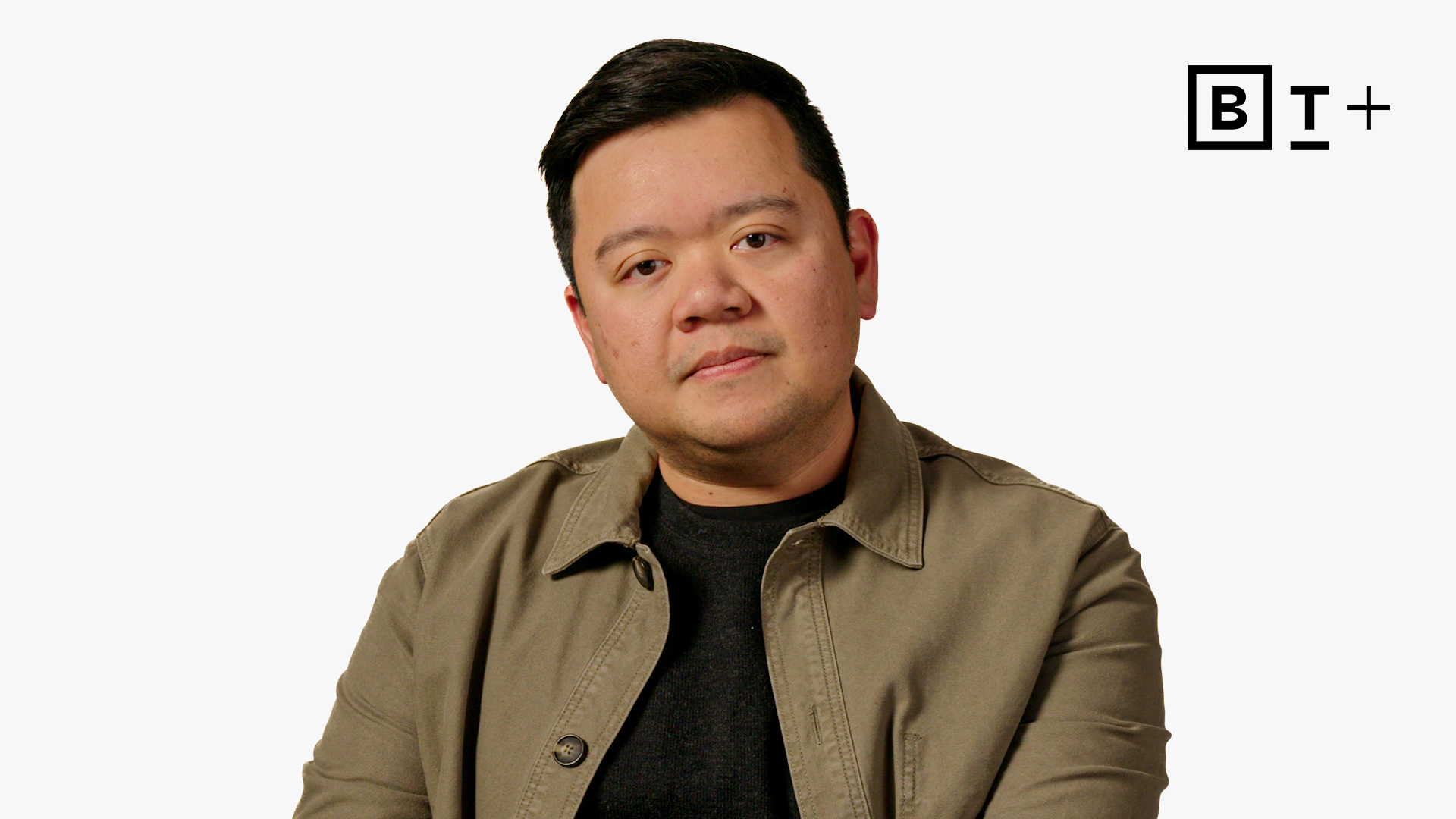 A man with short dark hair wearing a khaki jacket and black shirt sits against a plain white background with a "B T +" logo in the top right corner.