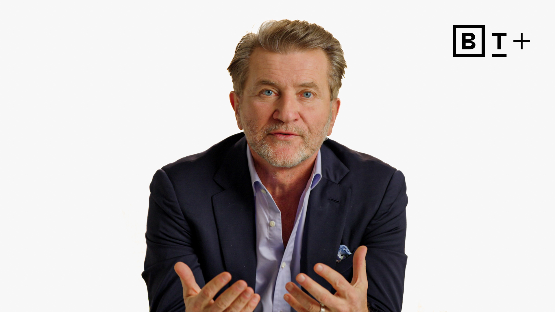 A man in a suit speaks with hands raised in front of a plain white background, next to a "B T plus" logo in the top right corner.
