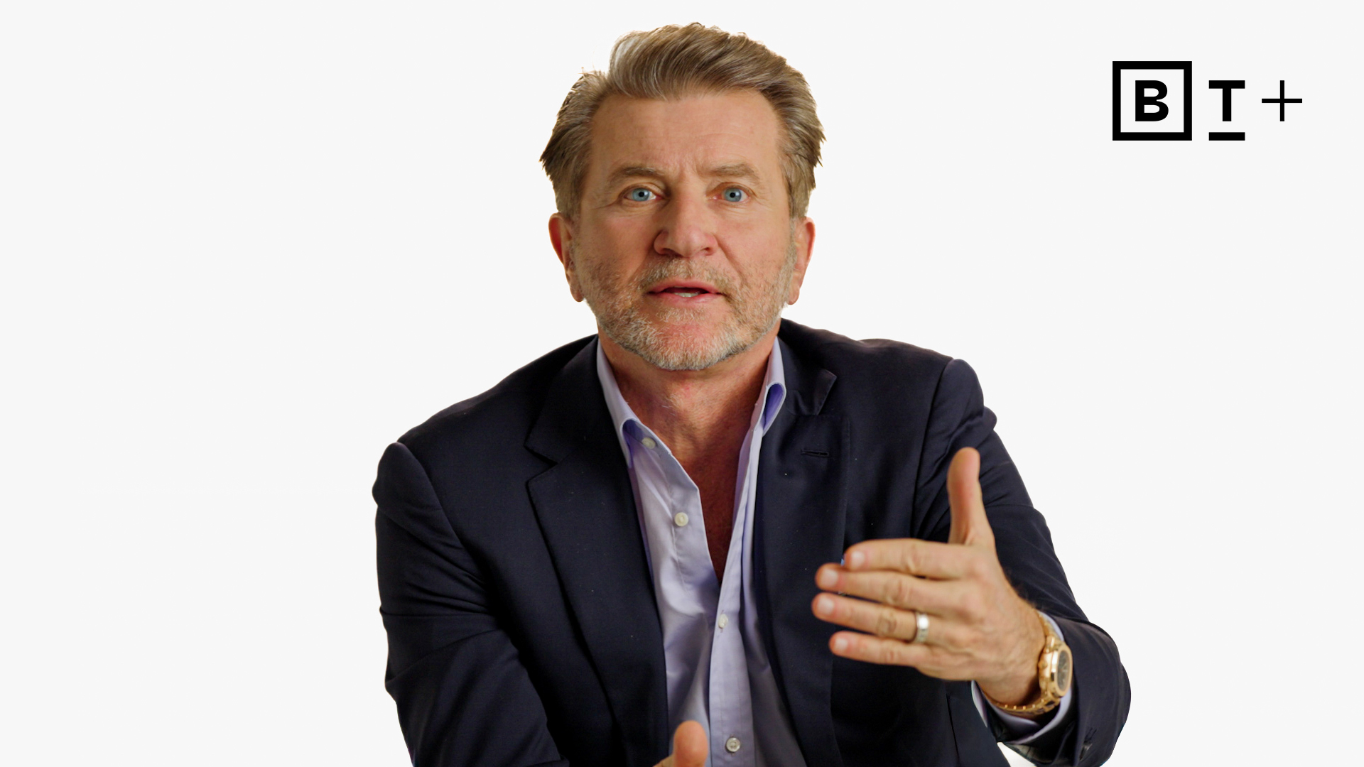 A man in a suit gestures with his hand while speaking, sitting against a plain white background with the B+ logo in the top right corner.