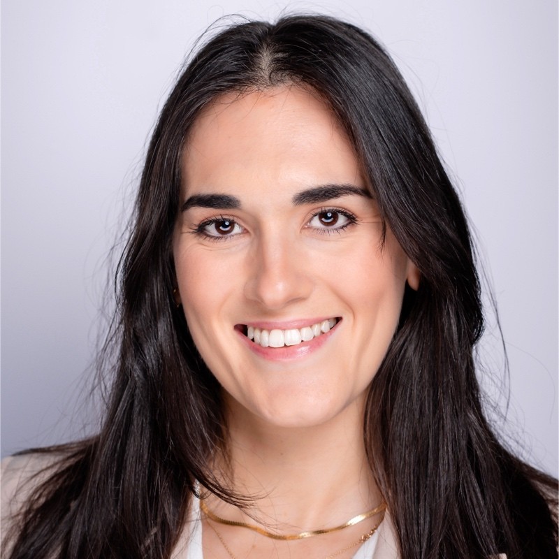 A woman with long dark hair smiles at the camera against a plain light background. She is wearing a light-colored top and a necklace.