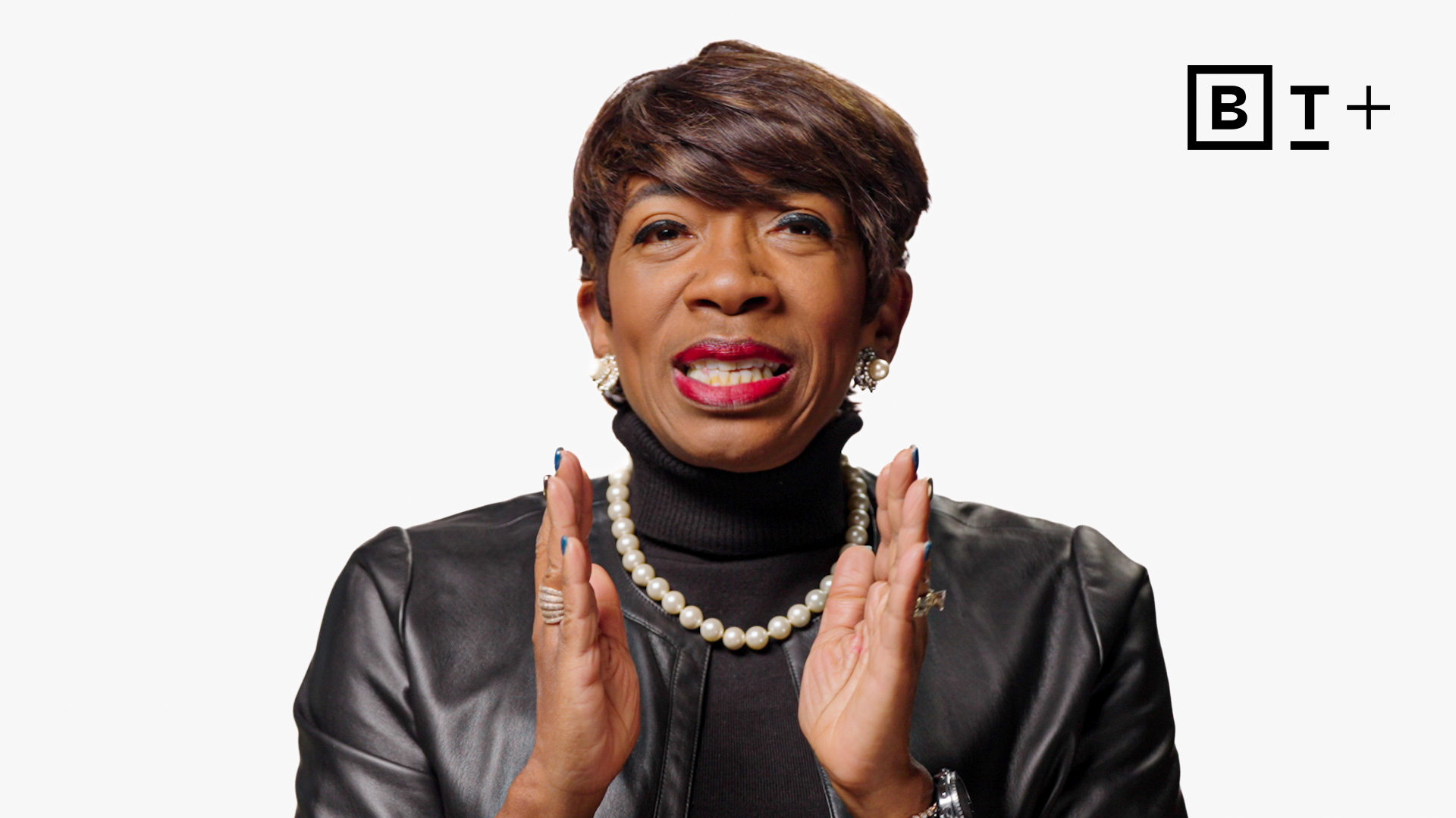 A woman in a black leather jacket and pearl necklace speaks with hands raised, against a white background with the B+ logo in the top right corner.