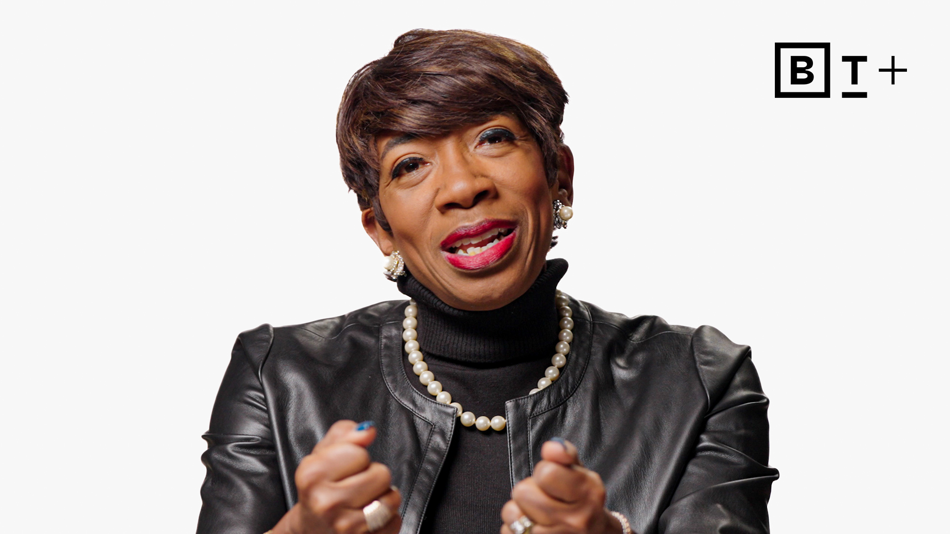 A woman in a black leather jacket and pearl necklace speaks while gesturing with her hands; BT+ logo is visible in the top right corner.