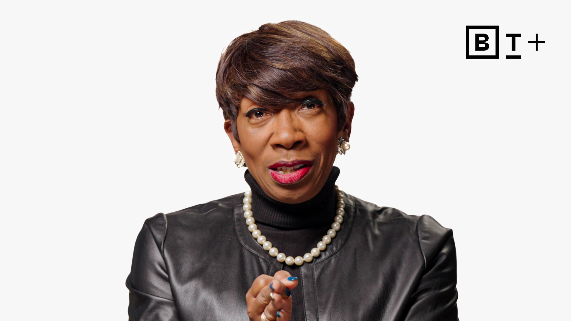 A person wearing a black leather jacket, black turtleneck, and pearl necklace speaks while gesturing, with a white background and a "B T +" logo in the top right corner.