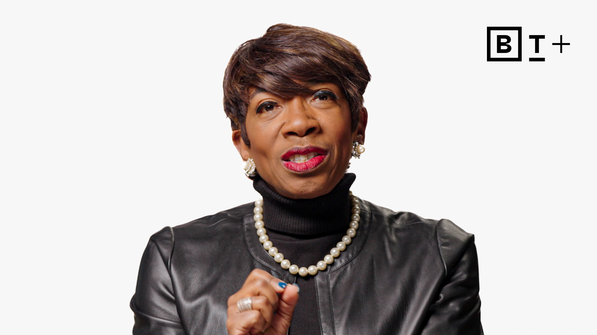 A woman in a black turtleneck and leather jacket speaks, wearing pearl earrings and necklace. A BT+ logo is visible in the top right corner on a white background.