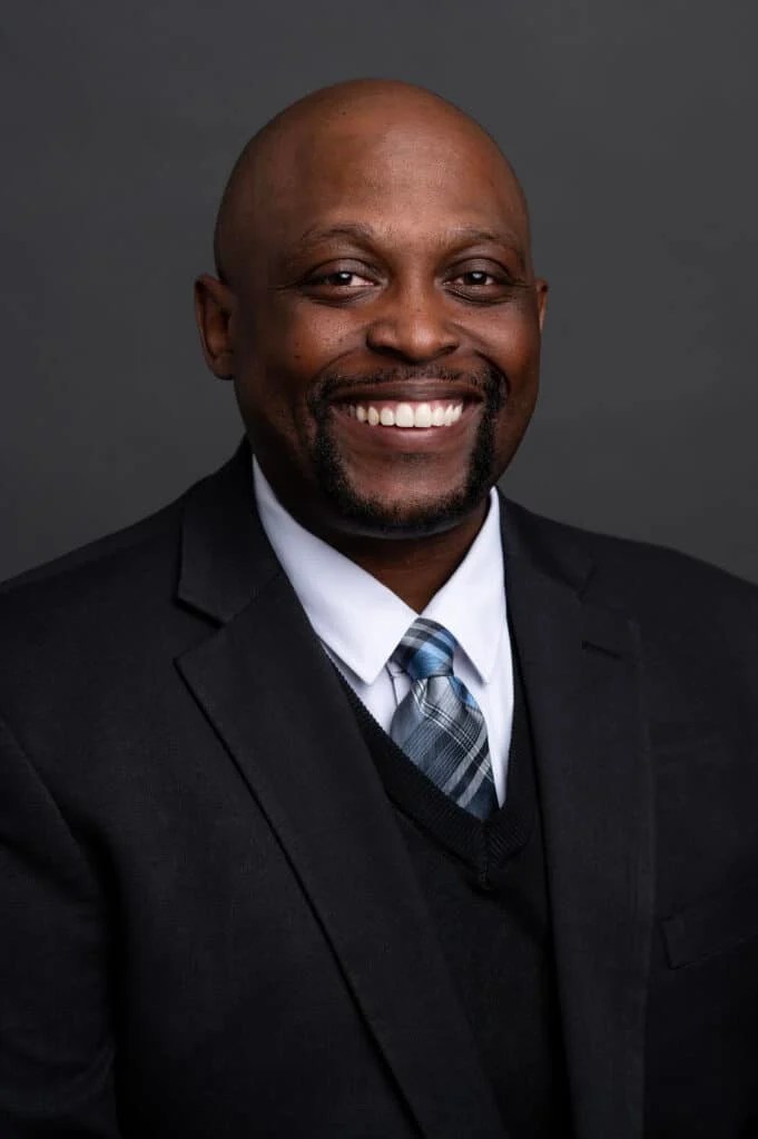 A man in a suit and tie smiles at the camera against a plain dark background.