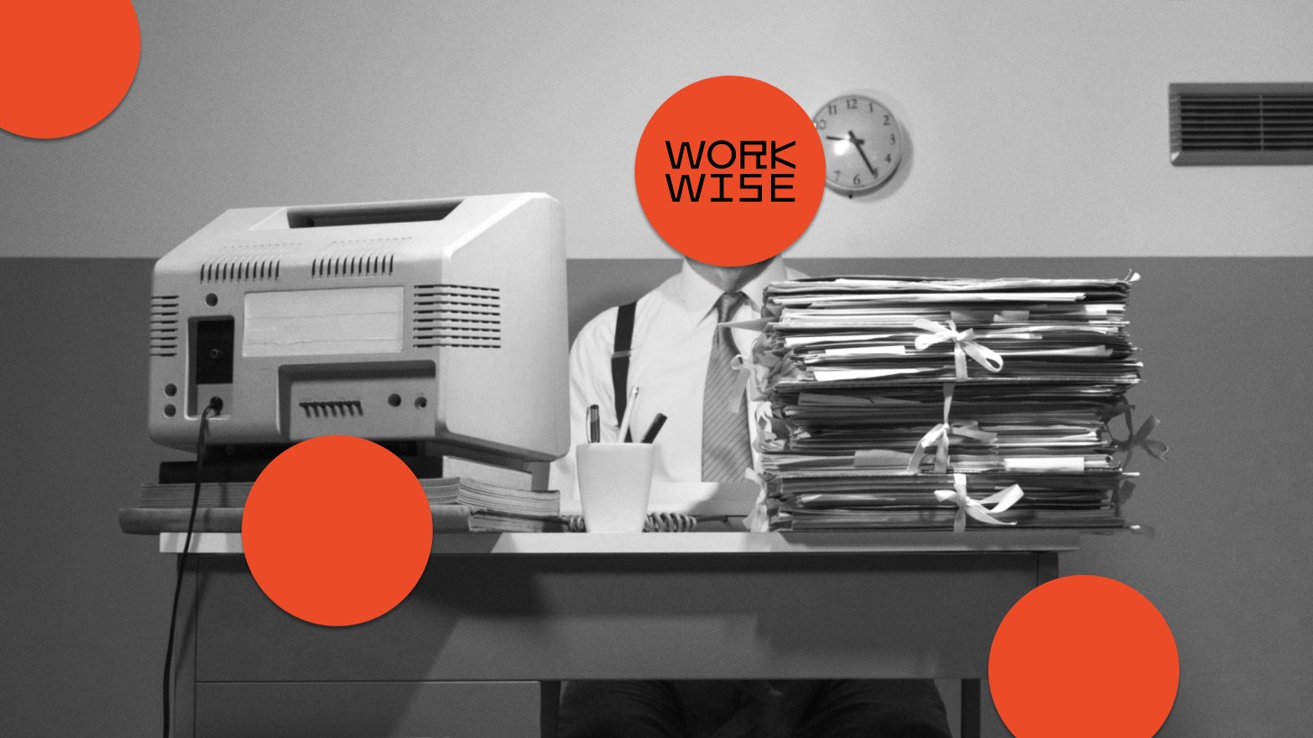 A person sits at a cluttered desk with a large stack of files, an old printer, and office supplies; their face is covered by an orange circle labeled "WORK WISE.