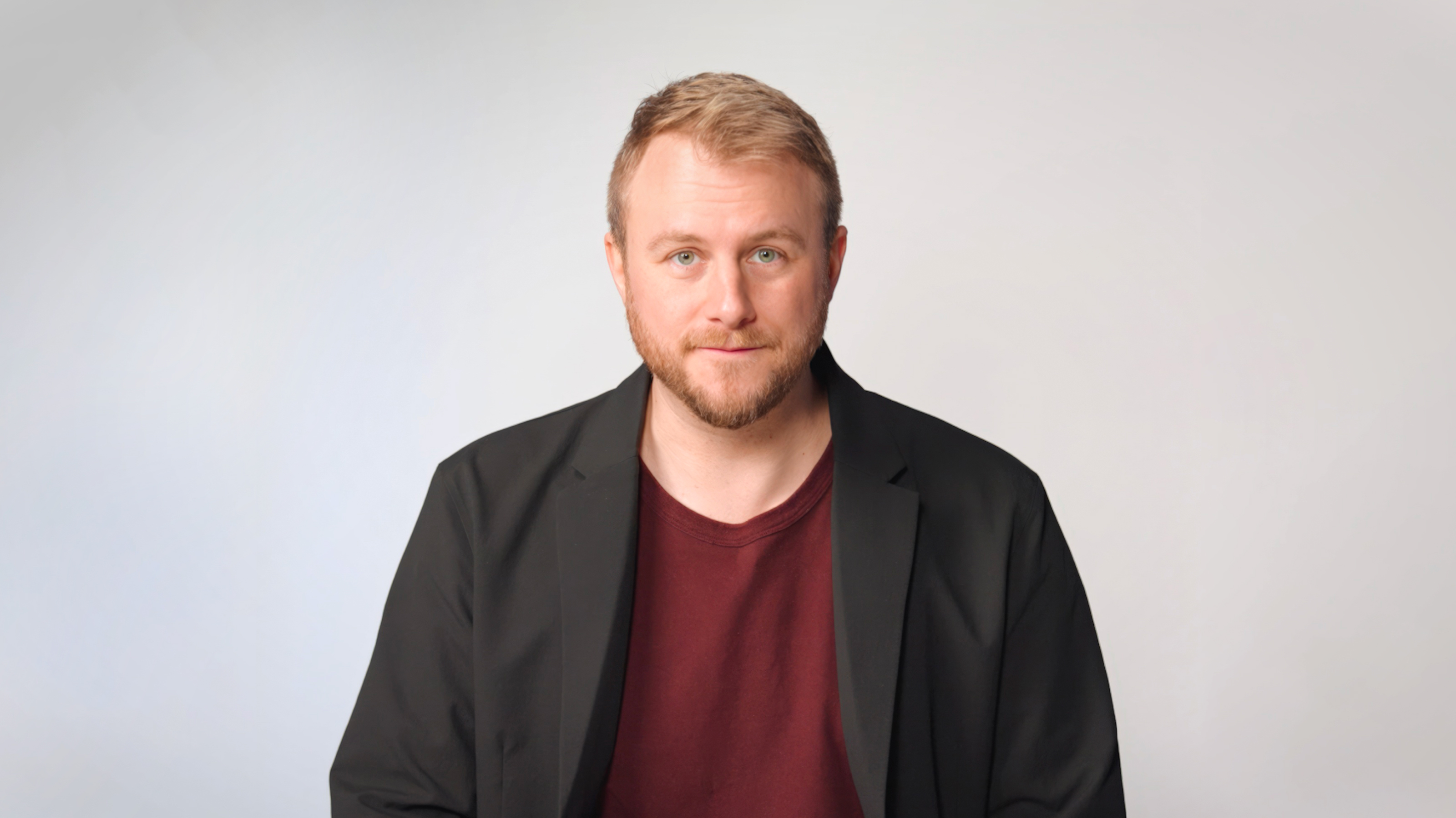A man with short blond hair and a beard wearing a black blazer over a maroon shirt sits against a plain light background, facing the camera.