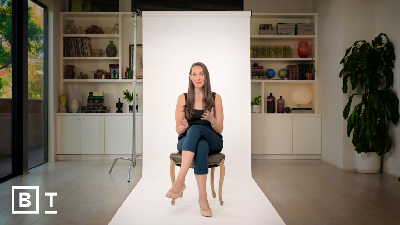 A woman sits on a chair in front of a white backdrop, gesturing with her hands. Shelves with books and decor are visible in the background. The BT logo is in the bottom left corner.