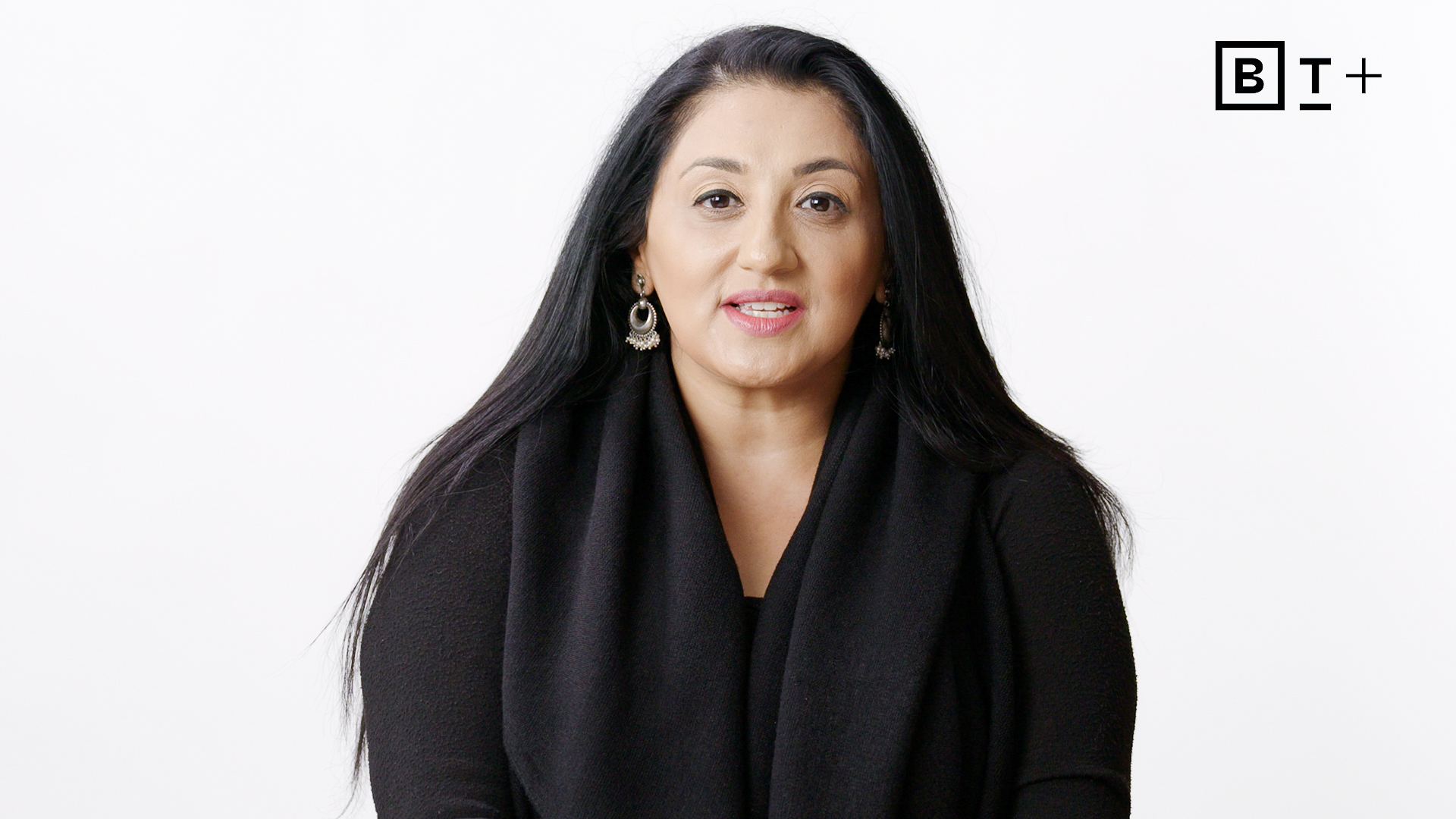 A woman with long black hair and earrings sits against a plain white background, wearing a black top. The B T Plus logo appears in the top right corner.