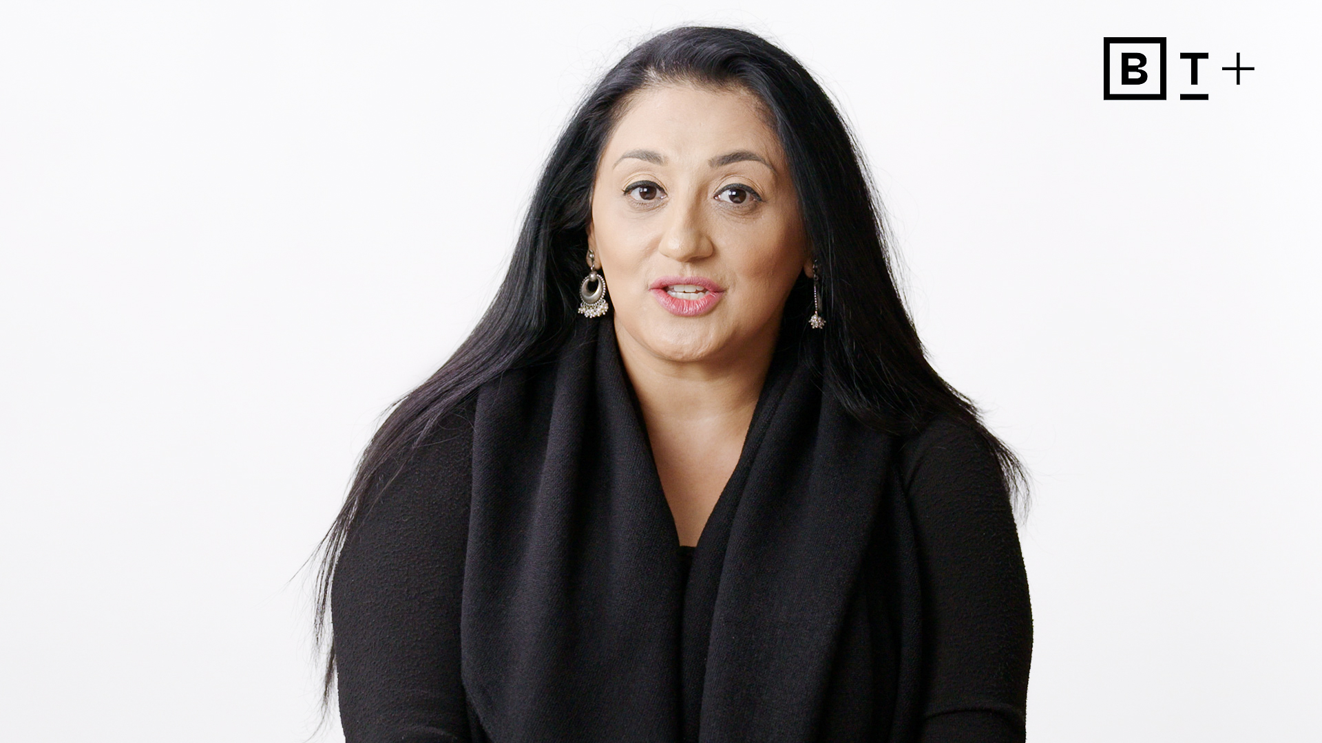A woman with long black hair, wearing a black top and earrings, speaks in front of a plain white background with the "BT+" logo in the top right corner.