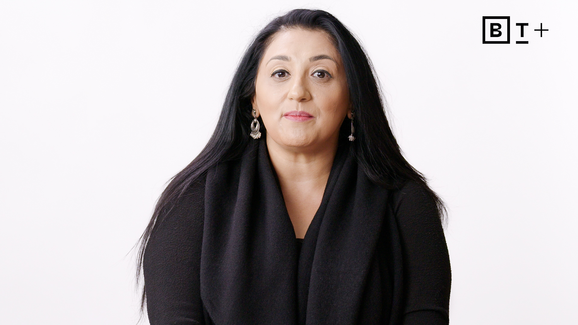 Woman with long dark hair wearing a black top and earrings sits in front of a plain white background. A "BT+" logo is visible in the top right corner.