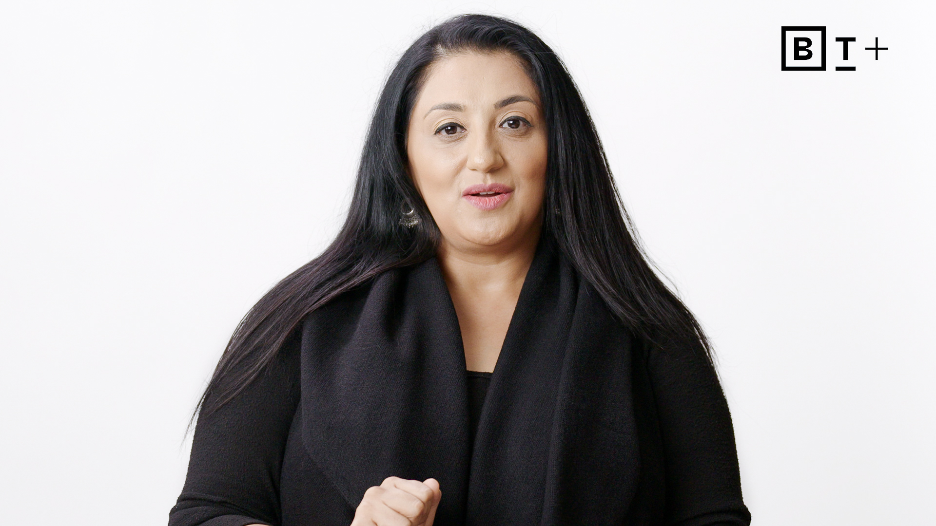 A woman with long dark hair wearing a black top stands in front of a plain white background with a "BT+" logo in the top right corner.