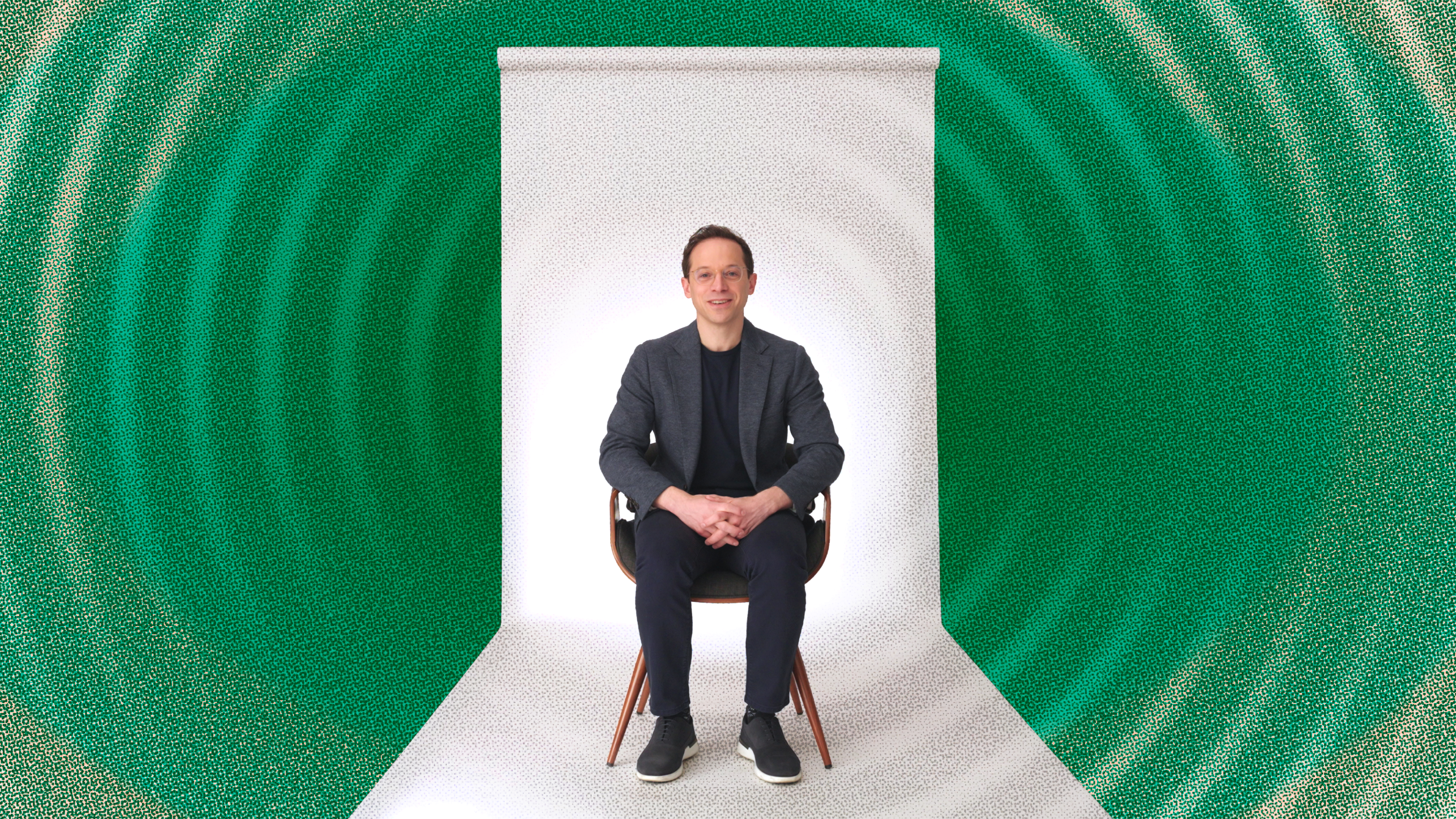 A man sits on a chair with hands folded in his lap, facing forward, against a white backdrop with green and teal concentric circles in the background.