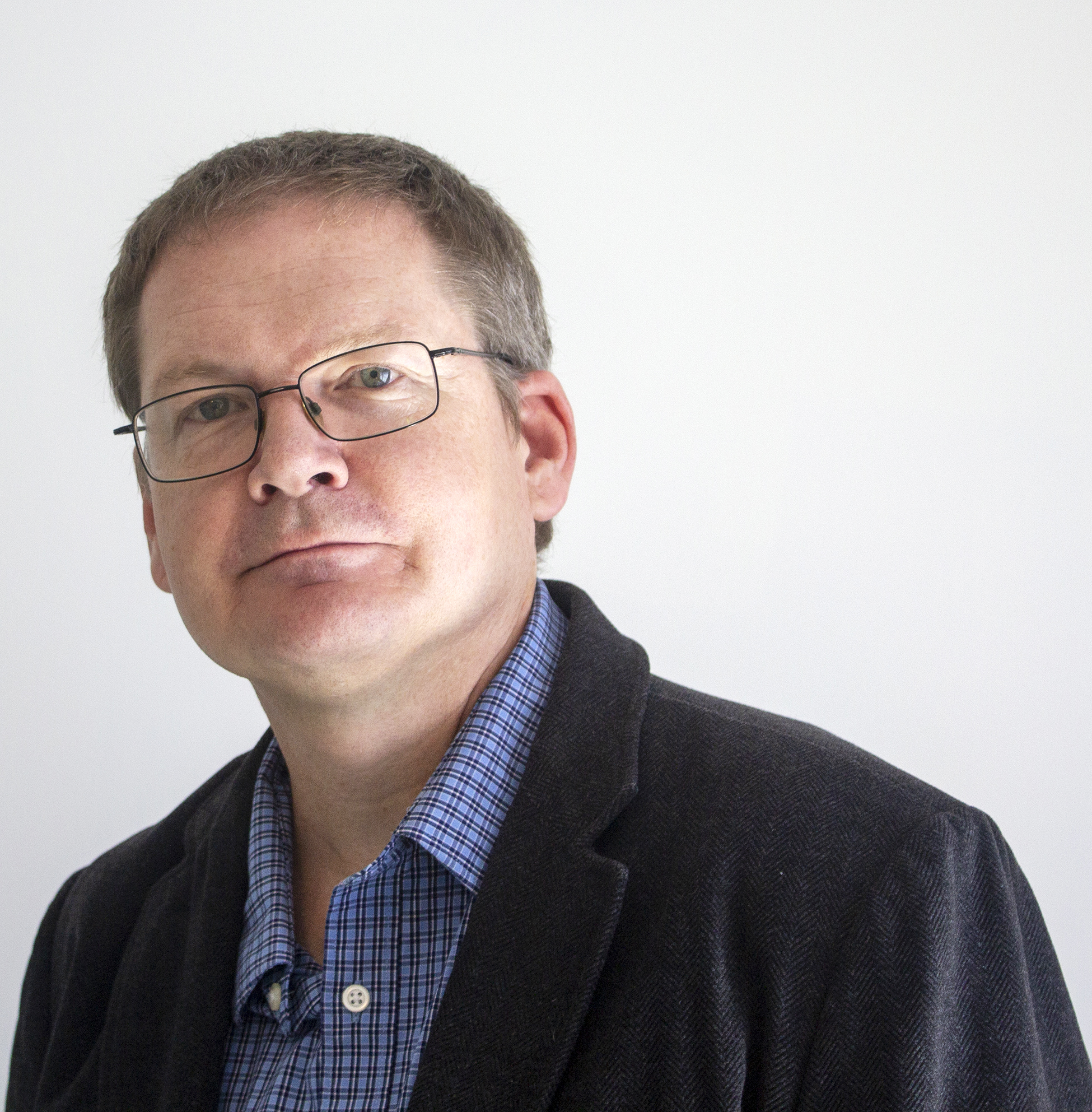 A middle-aged man with short hair and glasses wearing a blue checkered shirt and a dark blazer stands against a plain white background.