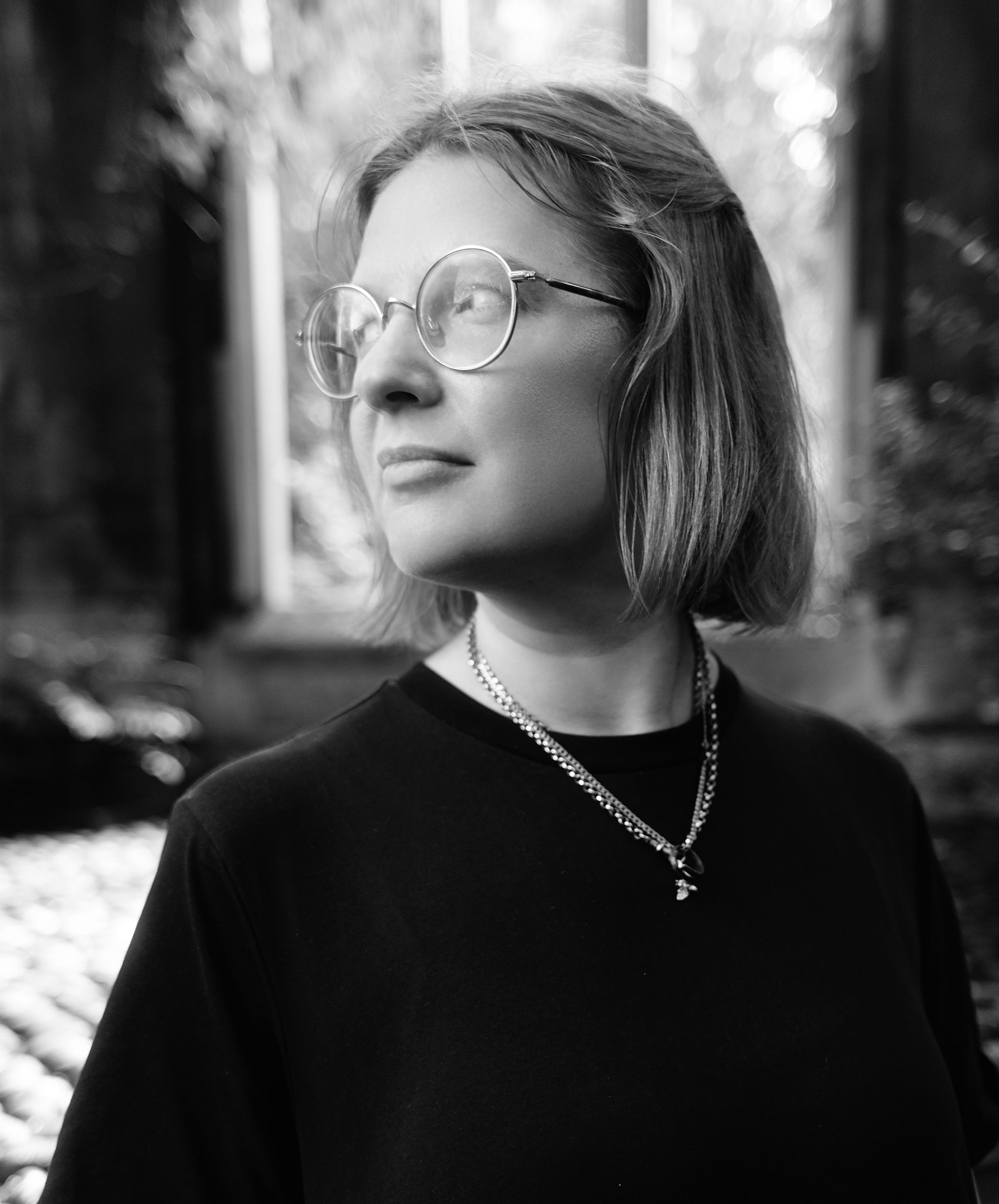 A person with short hair and glasses, wearing a dark t-shirt and necklace, stands indoors near a window, looking to the side. The image is in black and white.