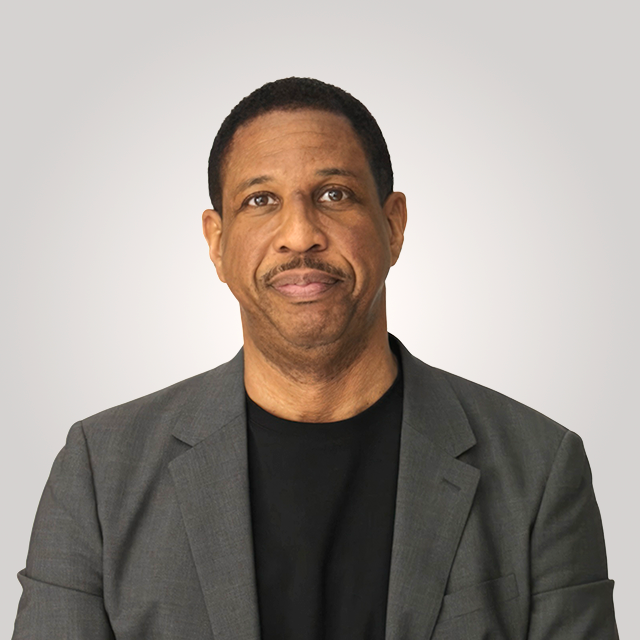 A man wearing a gray blazer over a black shirt stands against a plain light gray background, looking at the camera with a neutral expression.
