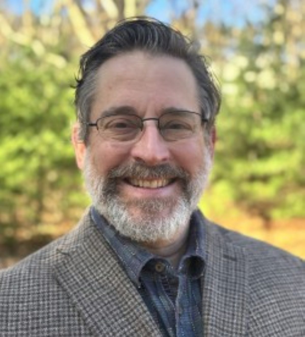 A middle-aged man with glasses, a beard, and graying hair, wearing a plaid blazer and collared shirt, smiles outdoors with trees in the background.