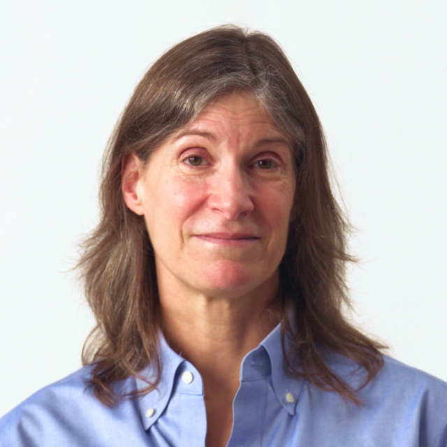 A middle-aged woman with shoulder-length brown hair wearing a light blue button-up shirt, facing the camera against a plain white background.