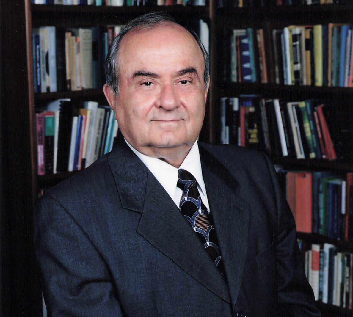 An older man in a dark suit sits in front of bookshelves filled with books, looking at the camera with a neutral expression.