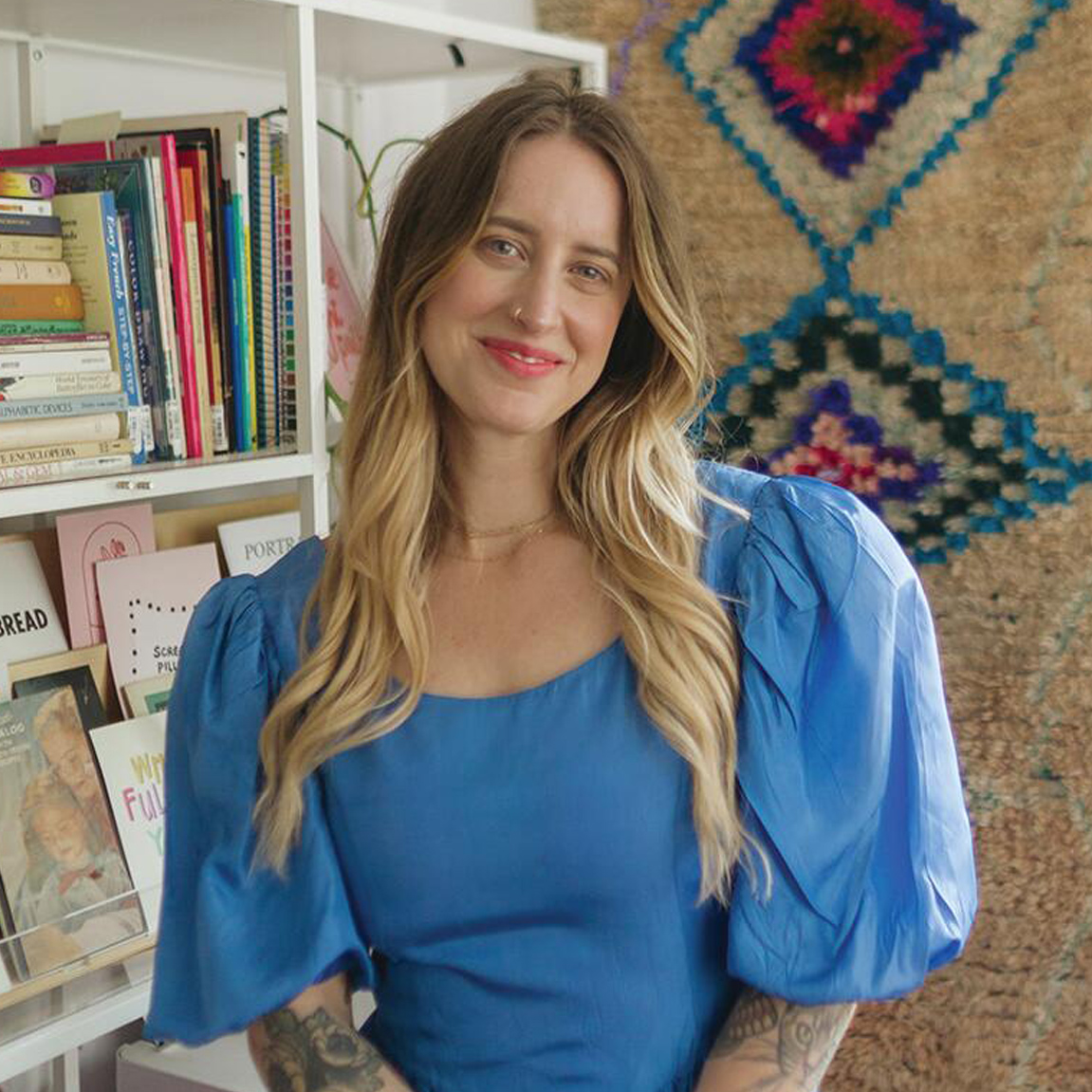 A woman with long blonde hair wearing a blue dress stands in front of bookshelves and a textured rug with geometric patterns.