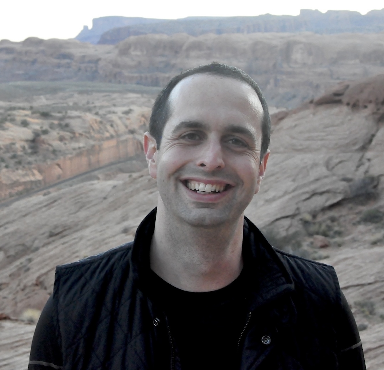 A man with short dark hair smiles at the camera, standing outdoors in a rocky desert landscape with mesas in the background.