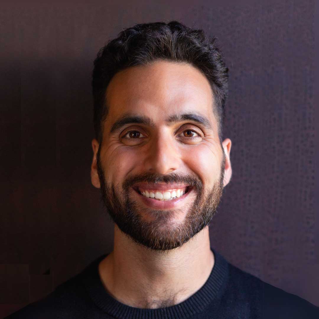 A man with dark hair and a beard is smiling at the camera. He is wearing a black shirt and is standing in front of a dark, plain background.